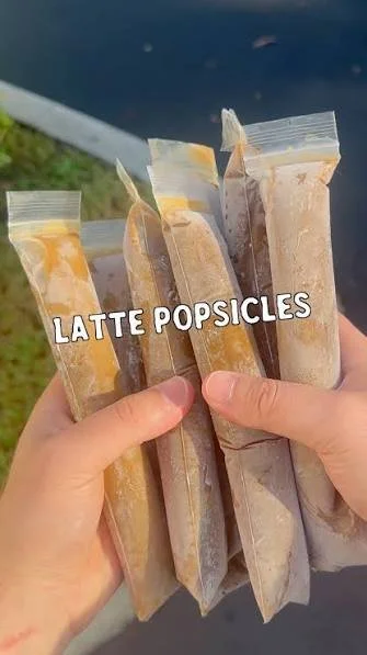 Hand holding five packaged latte popsicles outdoors, with the words "LATTE POPSICLES" overlayed.