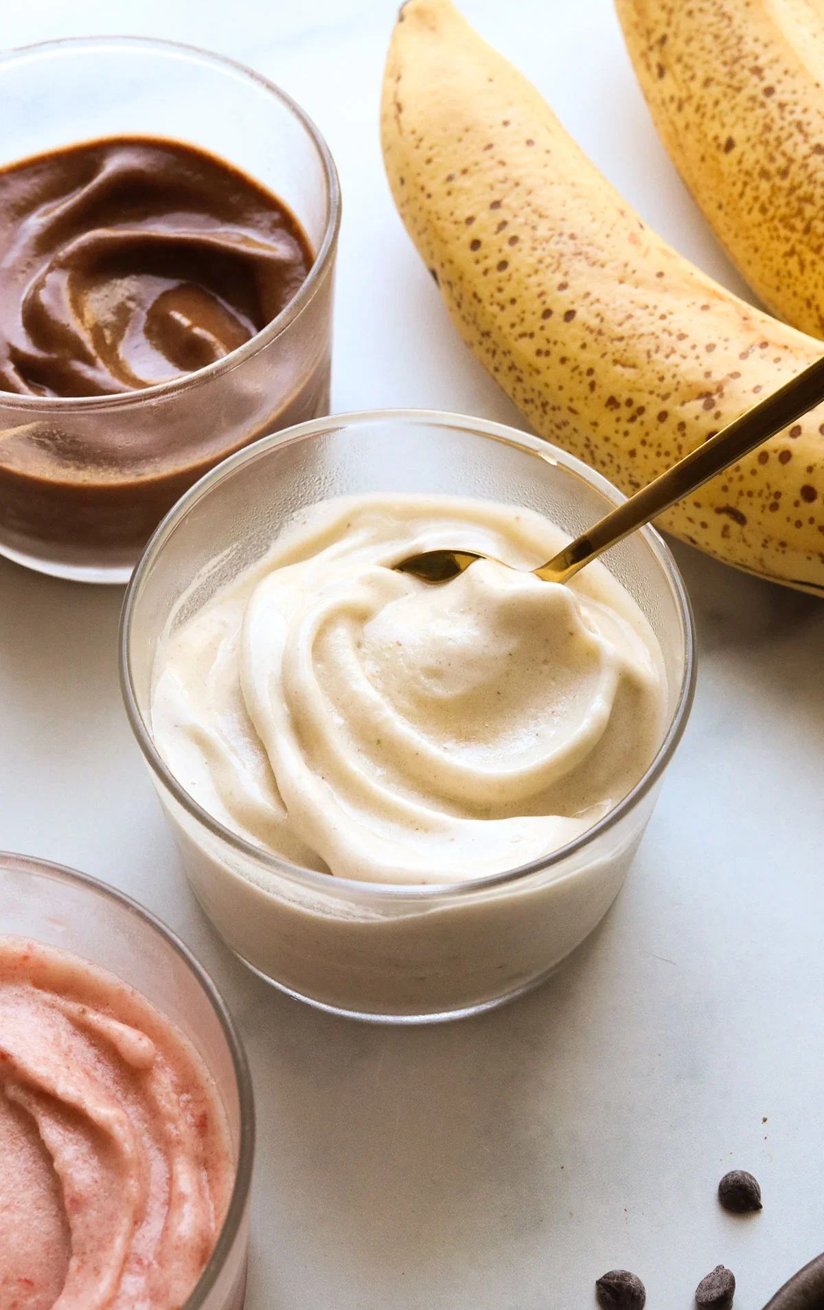 Two glasses of creamy dessert, one topped with a spoon, one with chocolate, next to a banana and small chocolate chips.