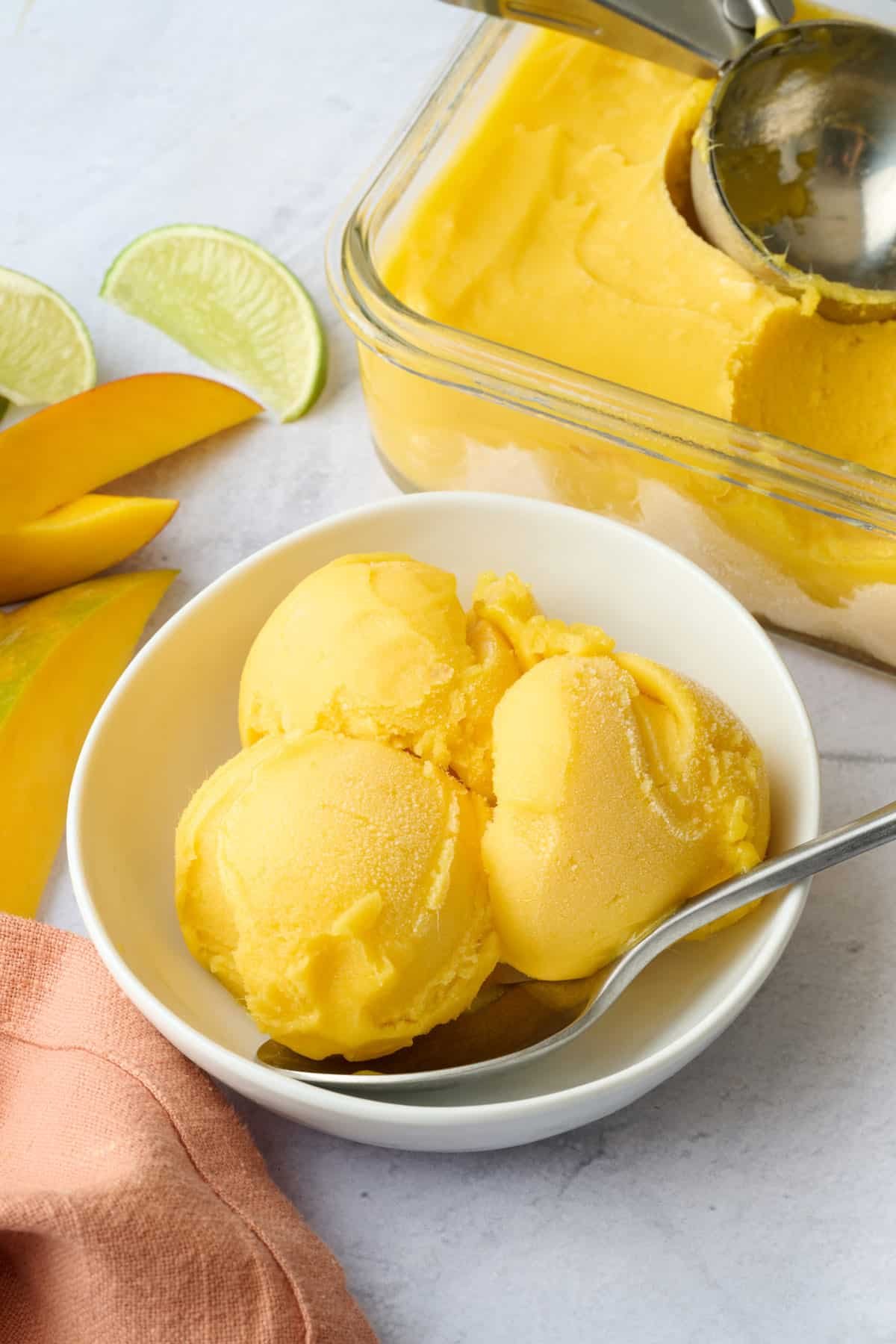 Three scoops of yellow mango ice cream in a white bowl on a marble surface, with a metal spoon. In the background, there is a glass container filled with mango ice cream and lemon and lime wedges, along with sliced mangoes.