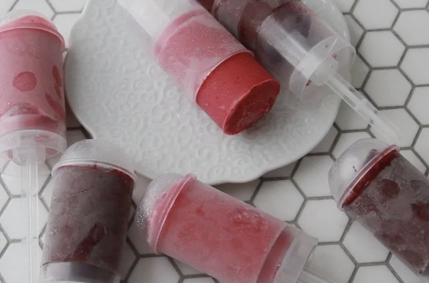 Six popsicles with red and purple fruit flavors on a white hexagonal tiled surface, some with foam or frost on them, on a white textured plate.