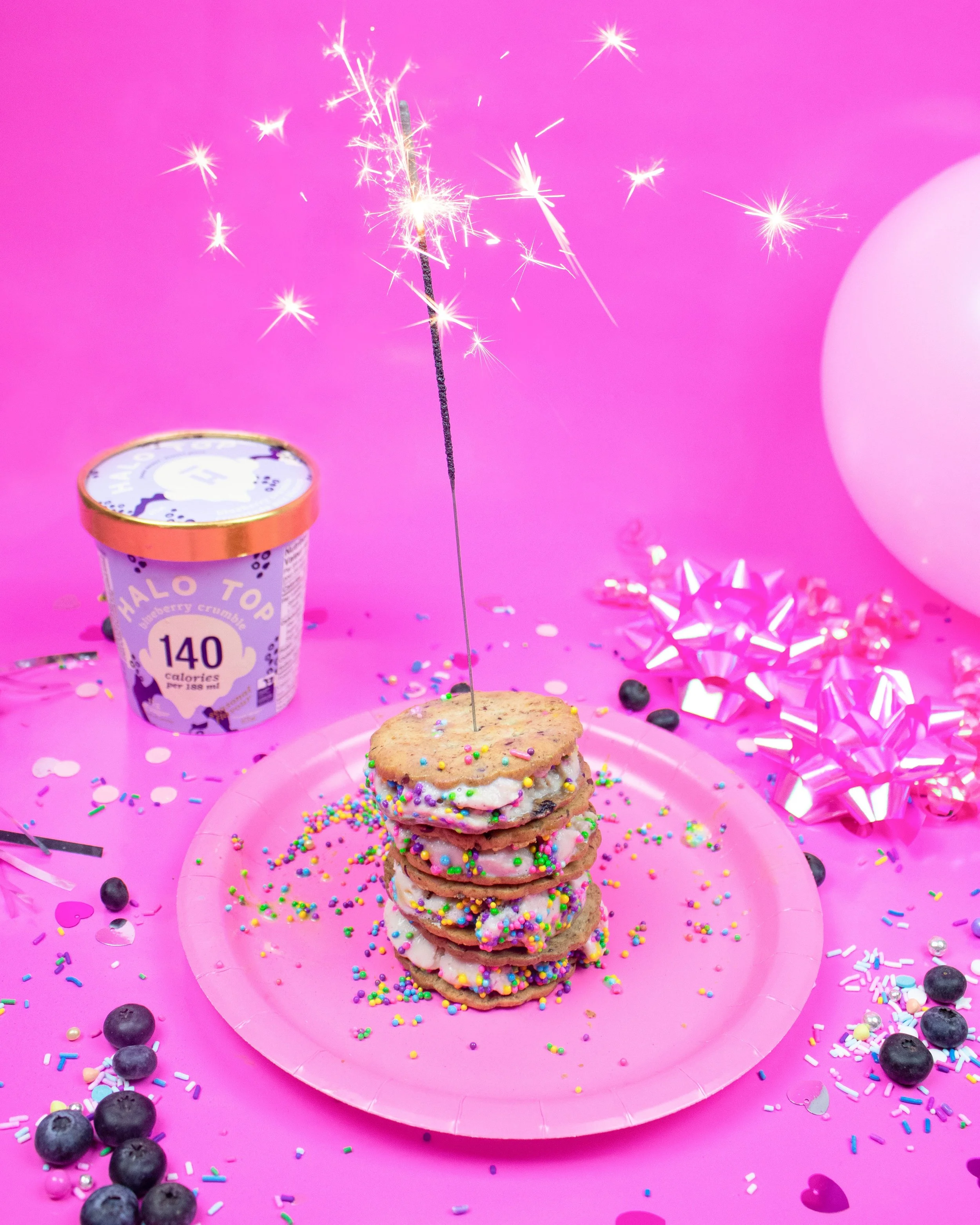 A stack of cookies with colorful sprinkles on a pink plate, surrounded by blueberries, confetti, and pink and silver ribbons, with a pink background and a pink balloon on the right. A container of Halo Top ice cream with a sparkler in front of it is also in the scene.