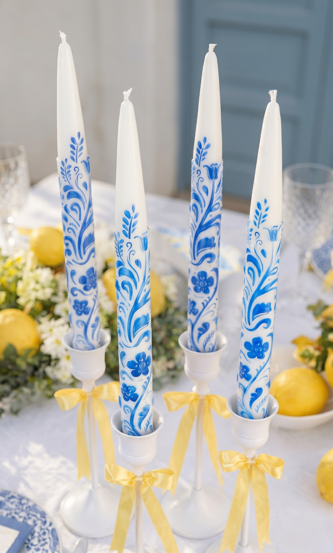 White taper candles with hand painted blue flowery decor on a white table with lemons and flowers