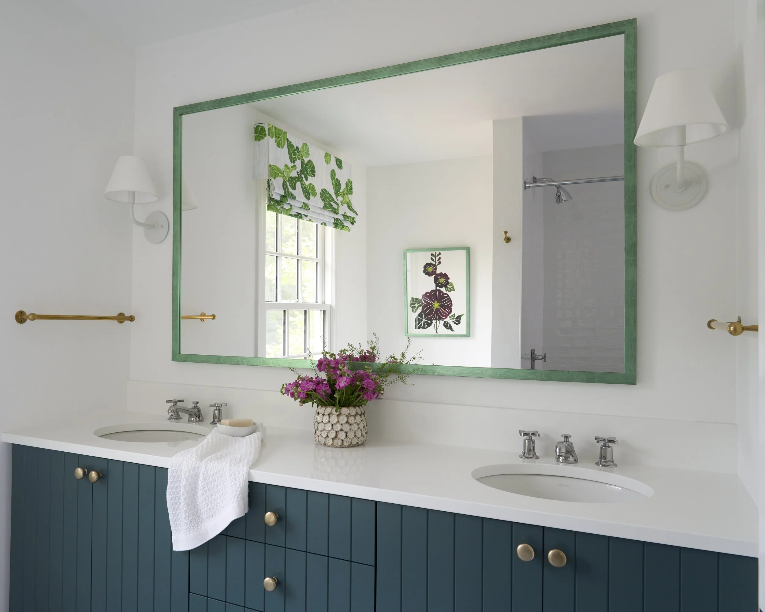 A bathroom vanity with a large mirror, a white countertop, two sinks, chrome faucets, blue cabinet with gold knobs, a white towel, a vase with pink flowers, a window with a green and white leafy valance, and framed floral artwork on the wall.