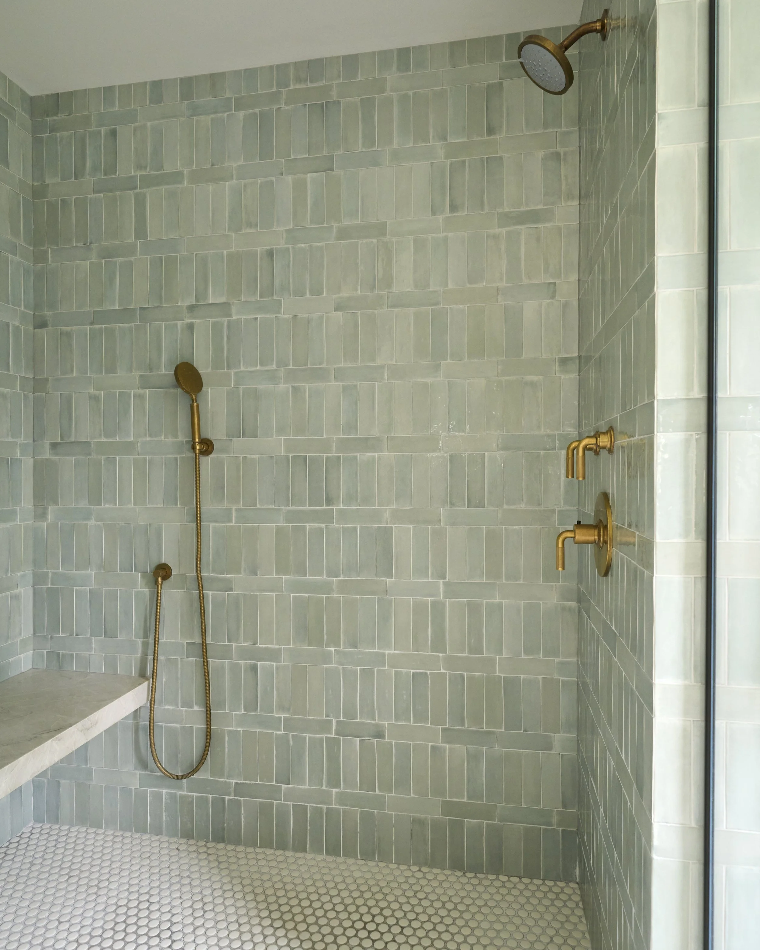 Shower area with green tiles, gold fixtures including a shower head, handheld shower, and controls, with a bench on the left side and a glass partition on the right.