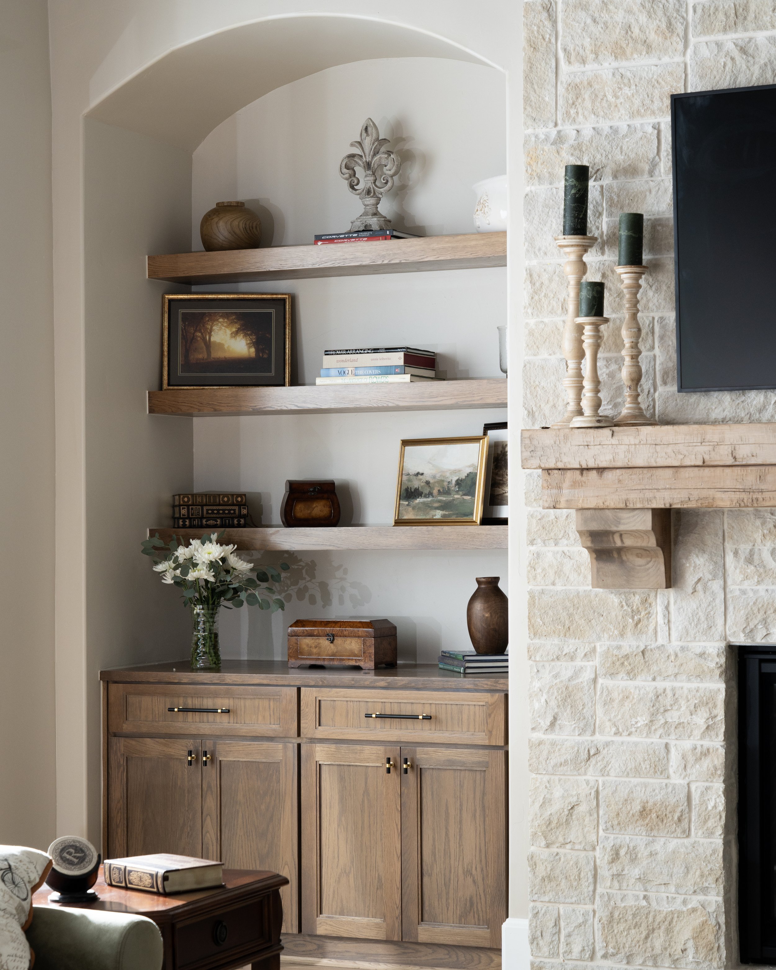 Interior view of a living room with wooden built-in shelves filled with decorative items, a picture frame, and books. On the right, there is a stone wall with three green candles on carved wooden candle holders. A wooden cabinet with doors is below, 
