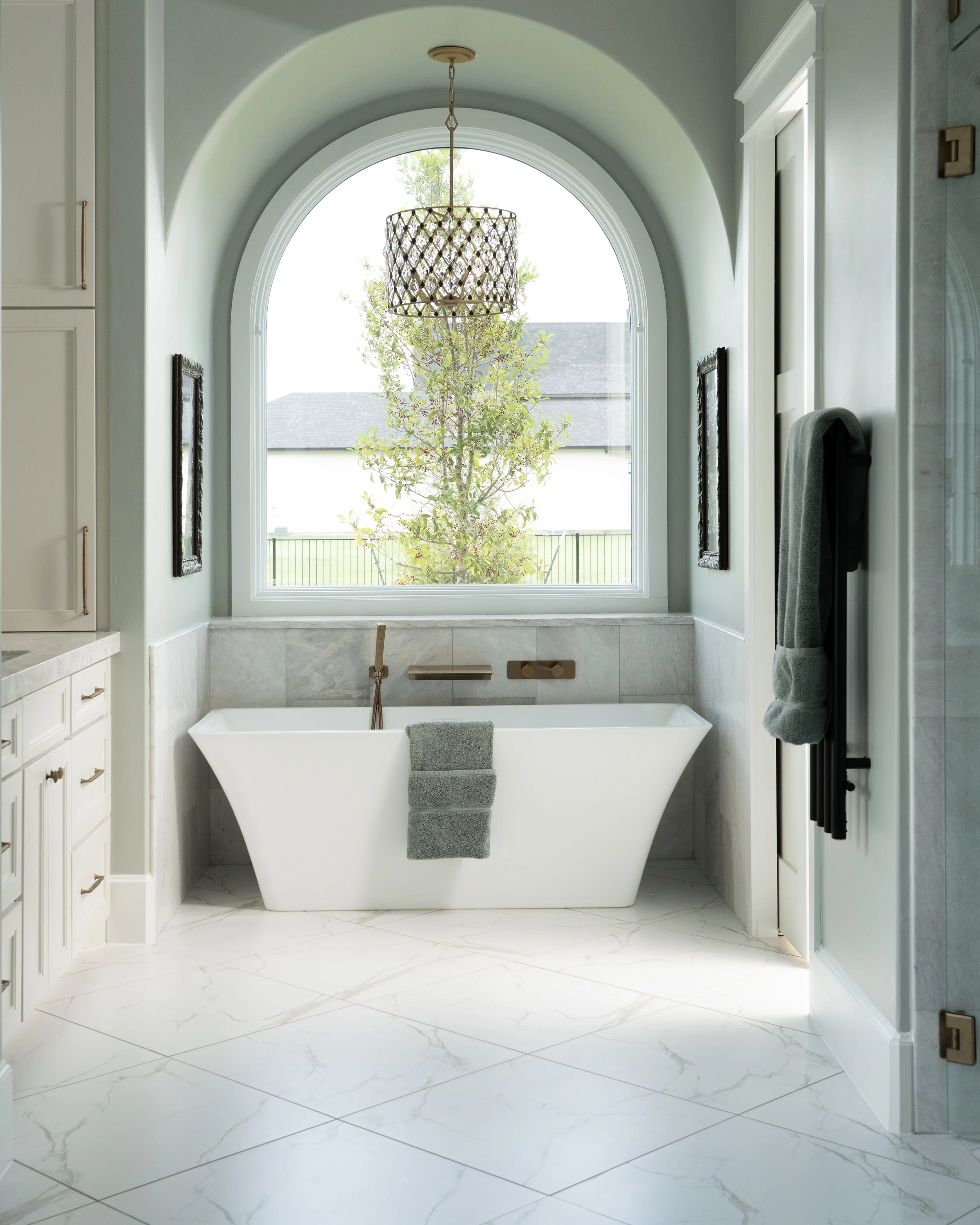 A bathroom with a white freestanding bathtub below a large arched window. Light green walls, marble flooring, and a chandelier hanging from the ceiling. A gray towel is draped over the edge of the tub, and a towel is hanging on a rack on the right si