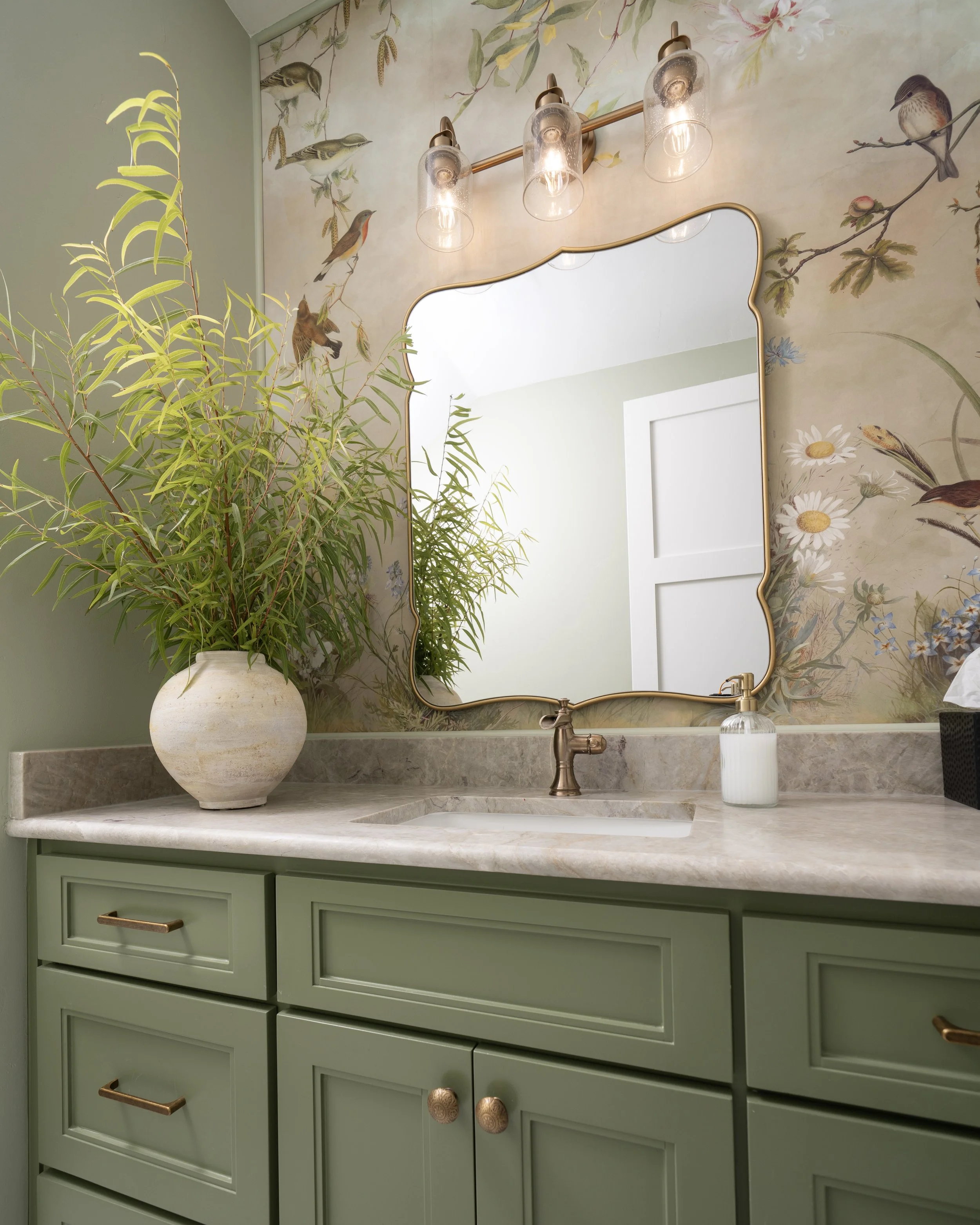 Bathroom vanity with a green cabinet, a marble countertop, a large mirror with a gold frame, a gold faucet, a white soap dispenser, a green plant in a round white vase, and a mural featuring various birds and flowers on the wall.