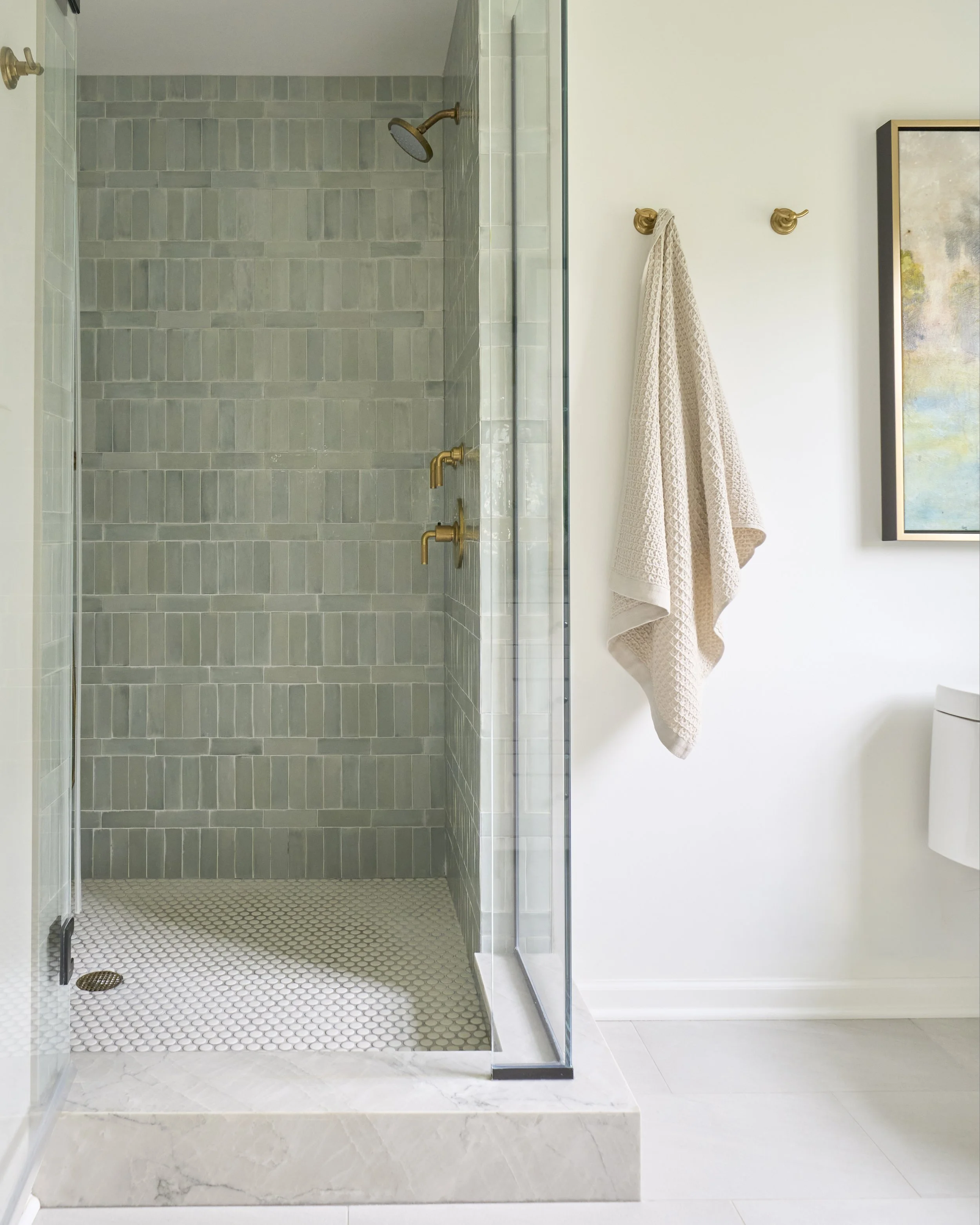 Modern bathroom with a glass-enclosed shower area featuring green tiles, a brass showerhead, and a matching brass faucet. The shower is on a raised marble platform, and there are gold hooks on the white wall to the right, holding a beige towel. A fra