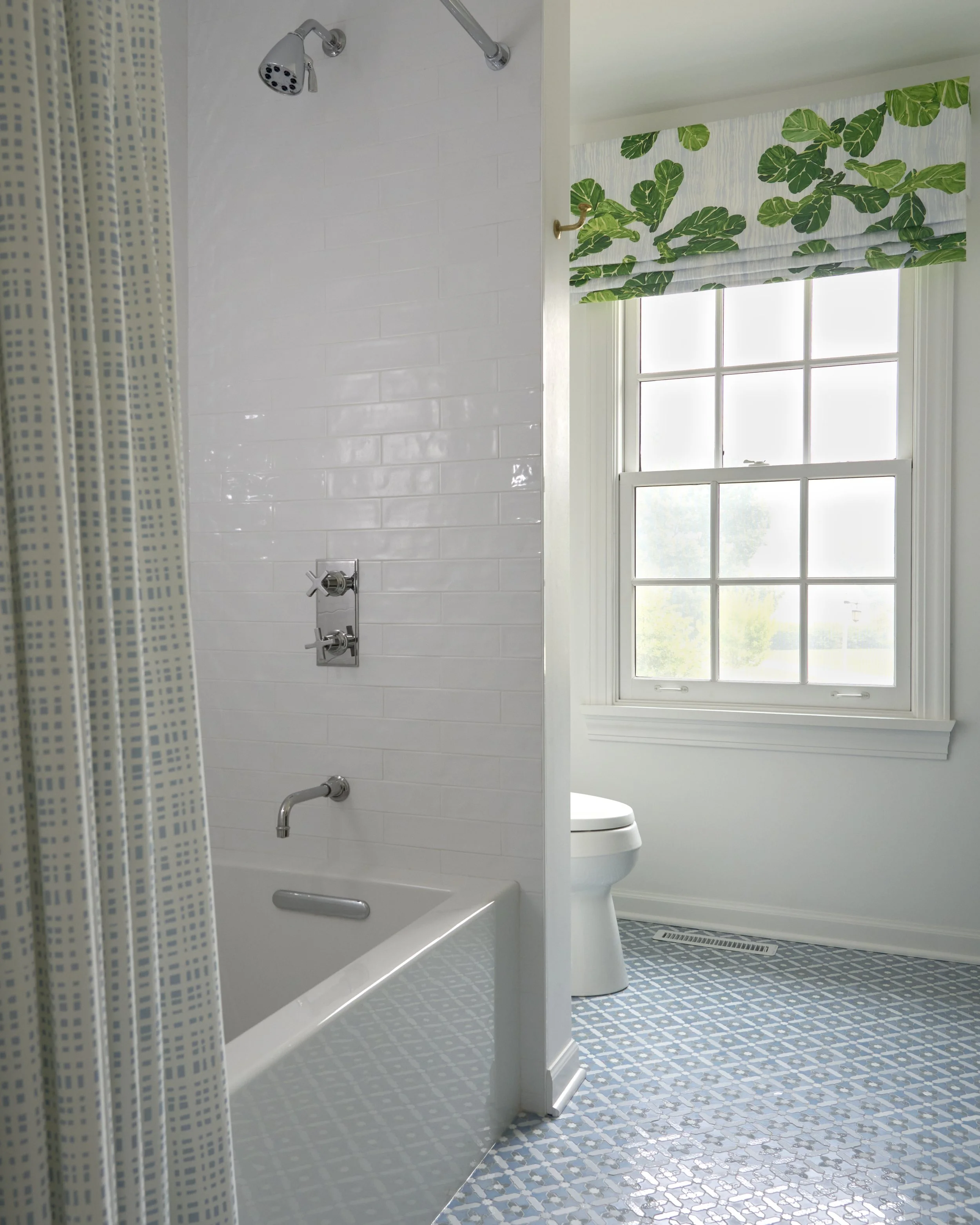 Bathroom with a white bathtub, gray patterned shower curtain, white tiled wall, window with green leaf-patterned Roman shade, and a white toilet visible in the background.