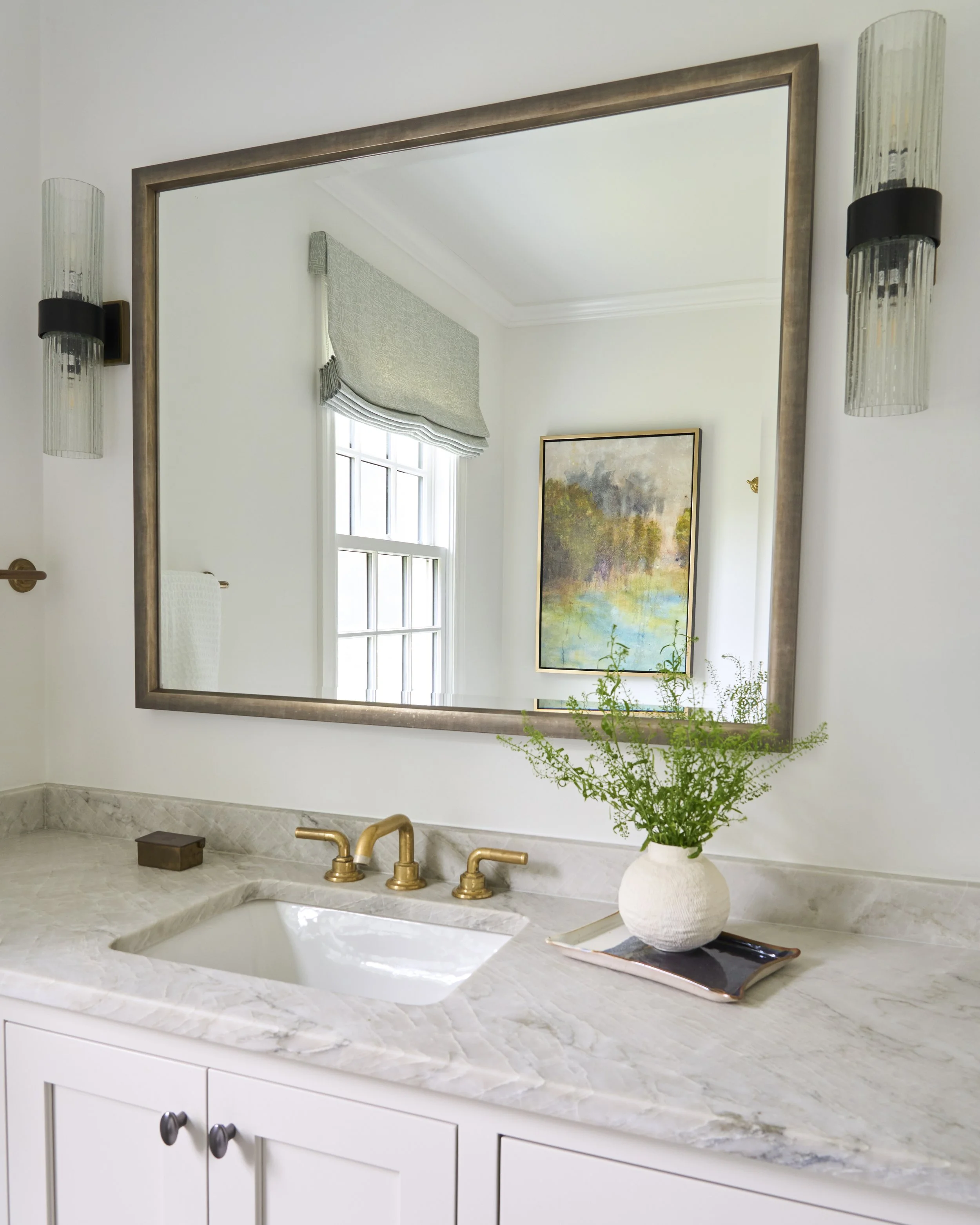 Bathroom vanity with a white marble countertop, gold faucet, decorative plant in a white vase, framed painting on the wall, mirror with a gold frame, and wall-mounted light fixtures, with a window and decorative window treatment located in Chicago