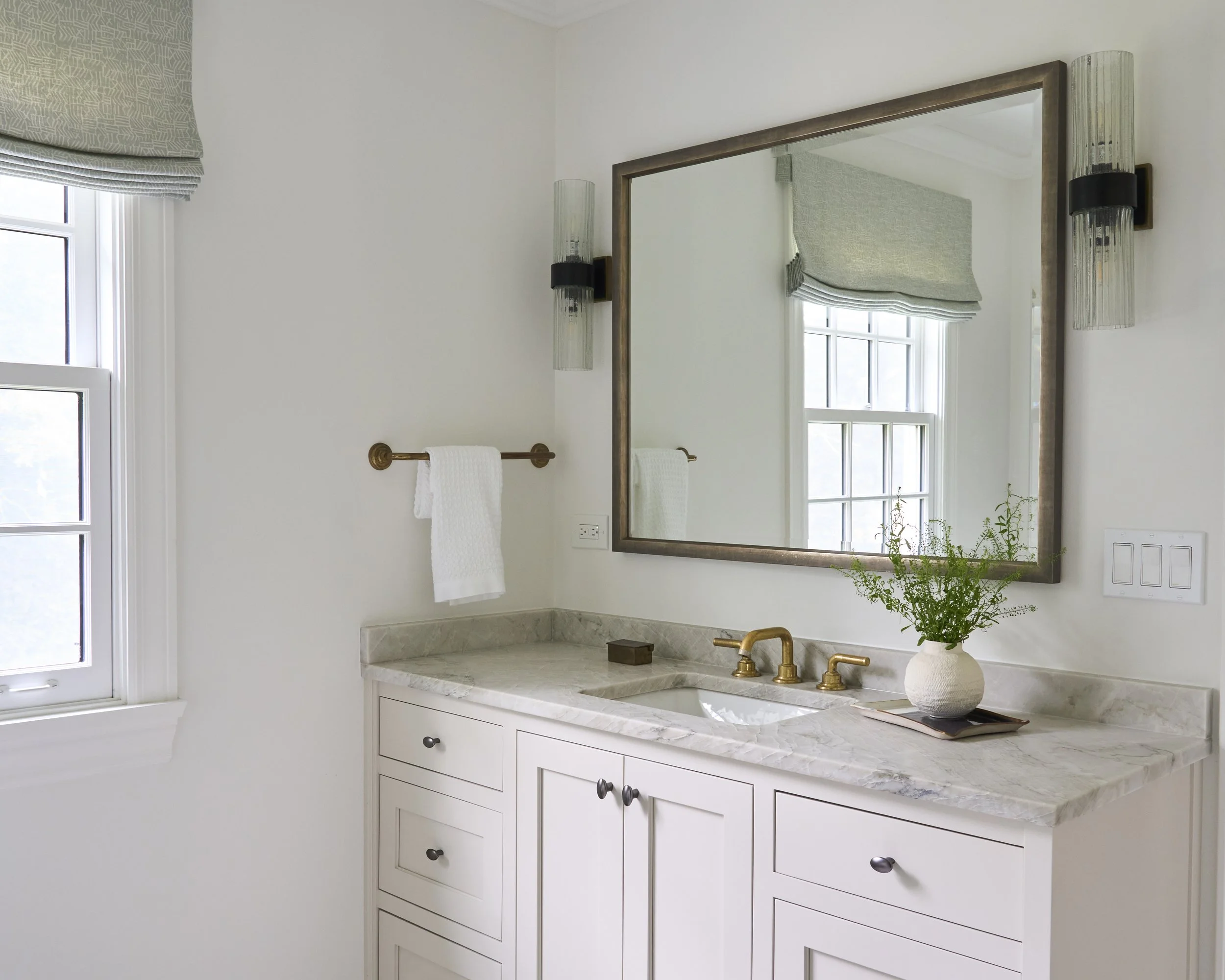 Bathroom vanity with a white cabinet, marble countertop, gold faucet, a tall mirror, wall-mounted lights, a window with a Roman shade, a towel rack with a white towel, and a potted plant.