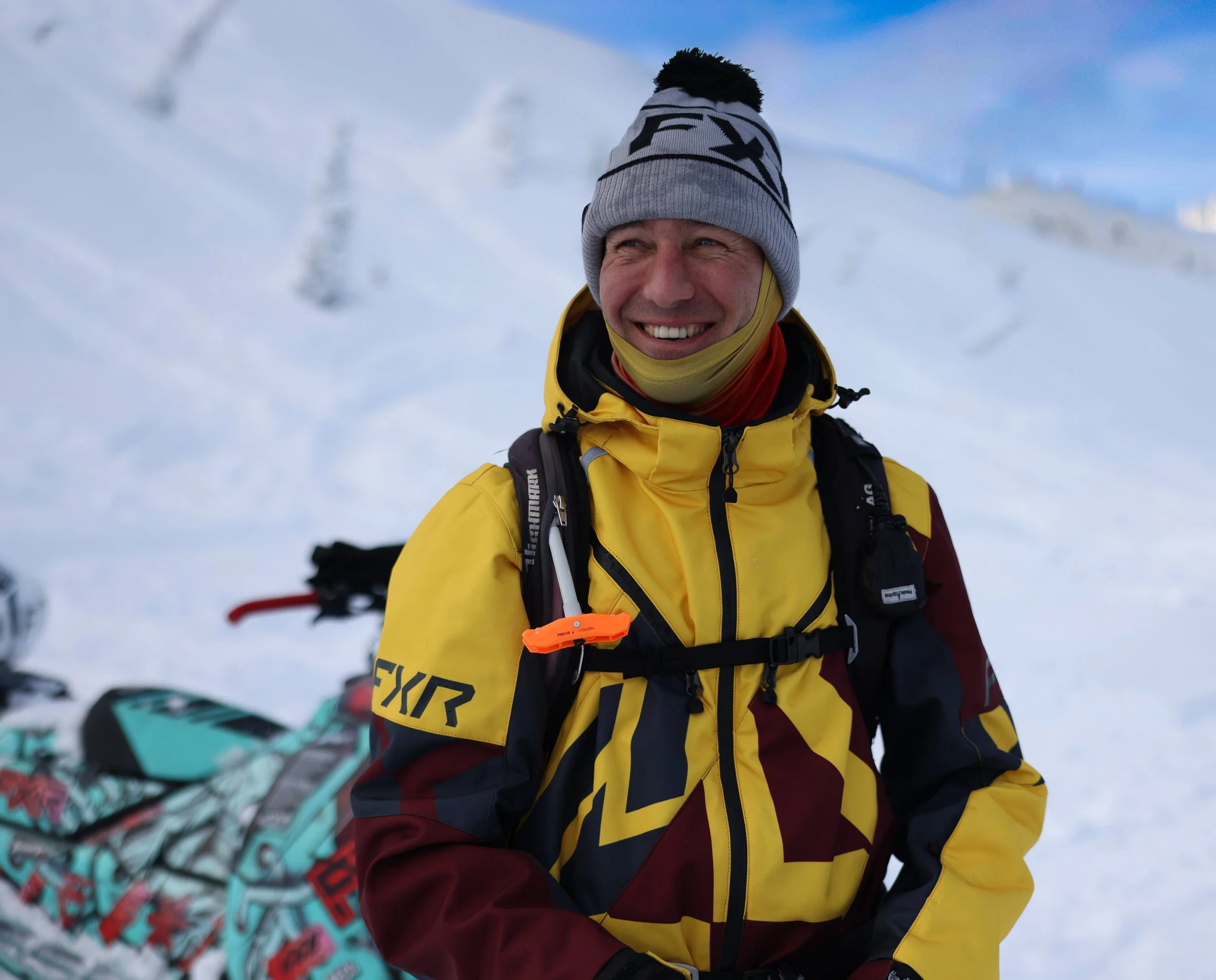 A man smiling outdoors in snowy conditions, wearing a yellow and maroon jacket, gray knit hat, and a neck gaiter, with snow-covered mountains in the background, and snowmobile nearby.