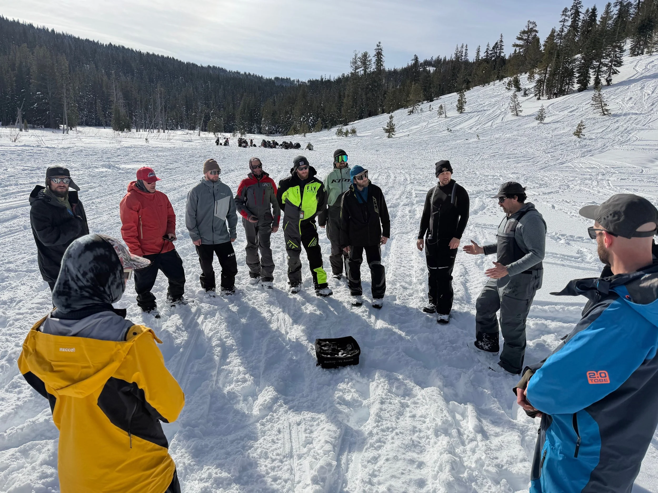 A group of people dressed in winter clothing gathered outdoors on snow-covered terrain, with snow covered mountains, engauged in avalanche education for snowmobilers