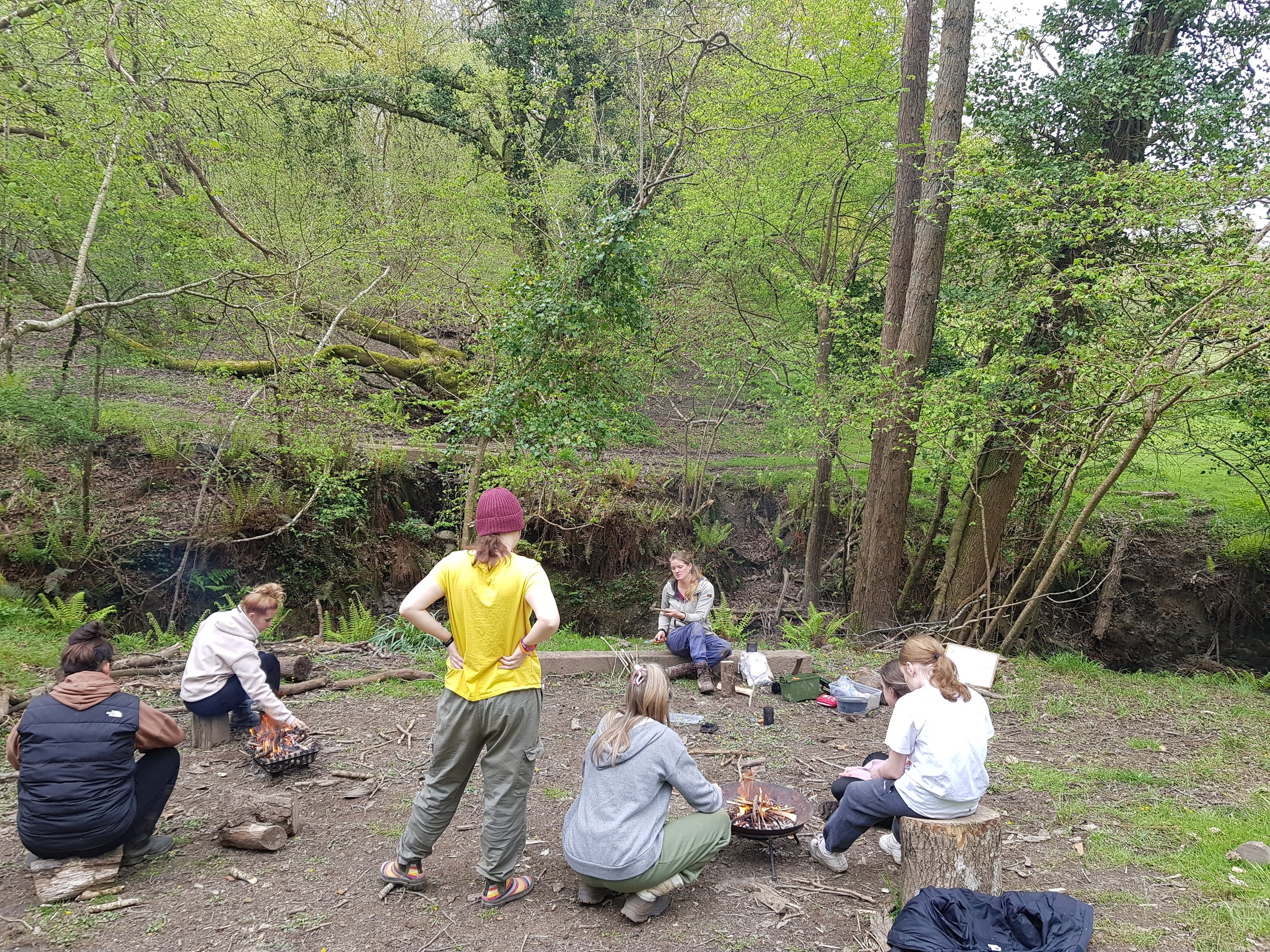 A group of children and teenagers gathered around small campfires in a forested area, with some sitting on logs and others standing, participating in an outdoor activity.