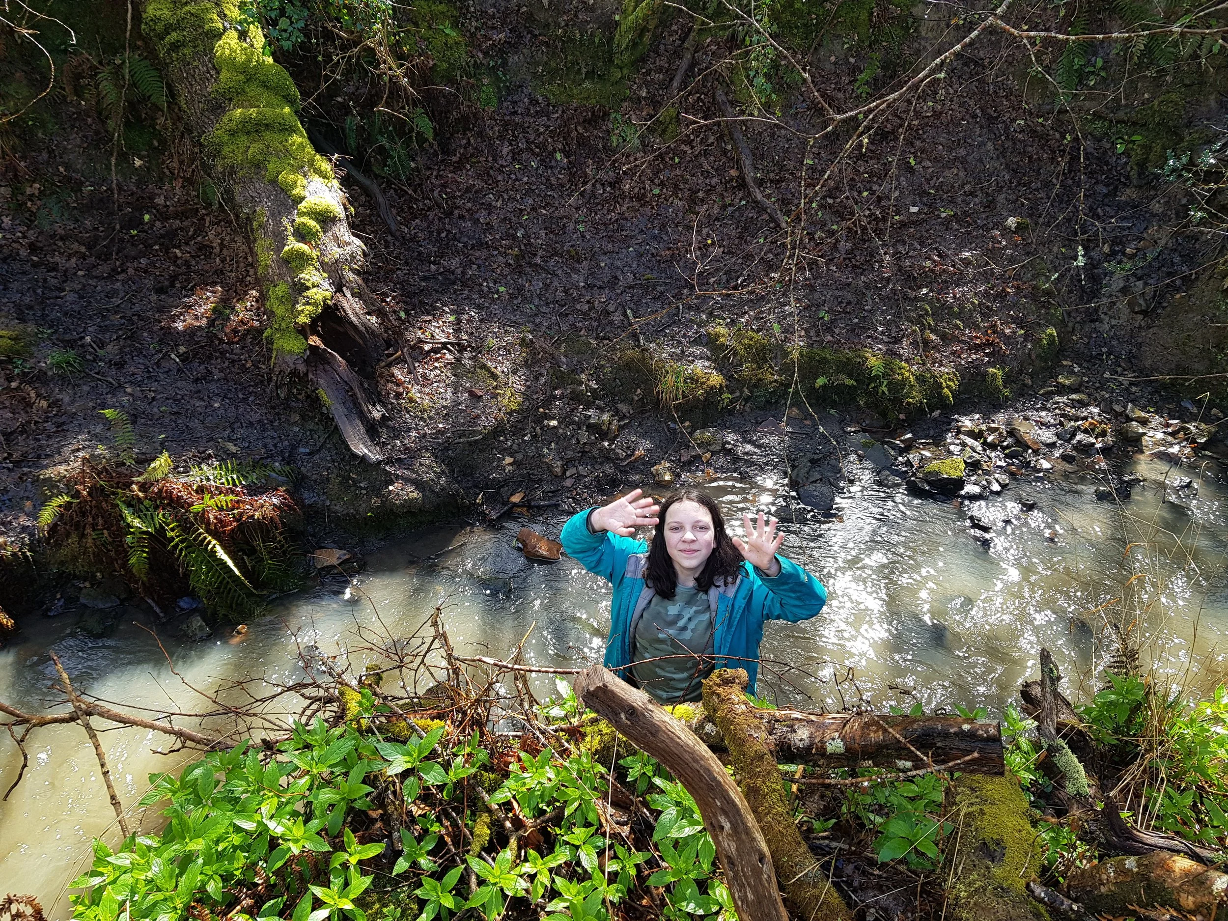 A young girl with long dark hair, wearing a camo shirt and a blue jacket, standing in a shallow creek surrounded by green plants and mossy logs, waving her hand and smiling up to the camera.