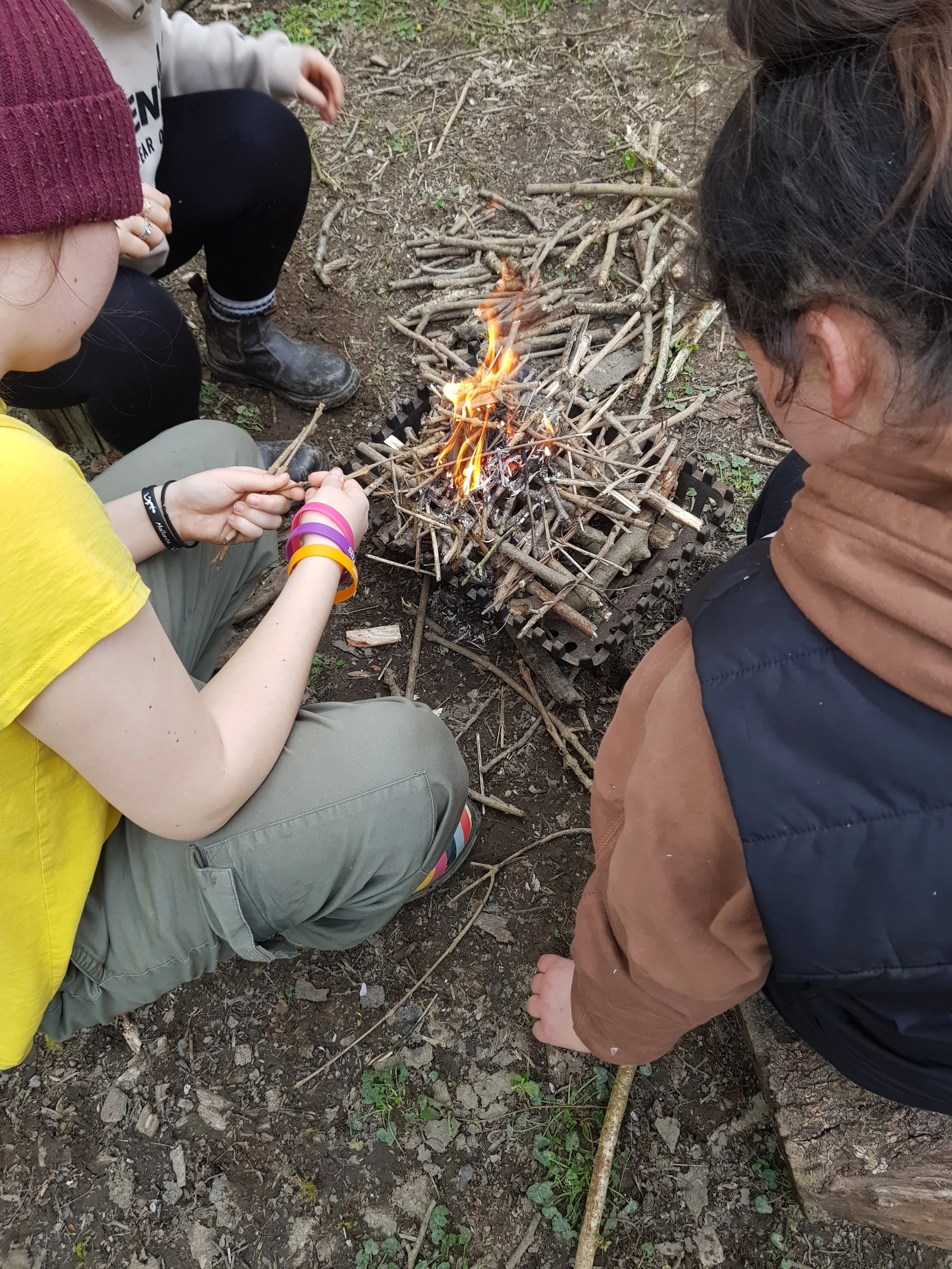 Three girls are sitting on dirt ground around a small campfire built with sticks, in an outdoor setting. One child is holding a stick over the fire, and the other two are watching.