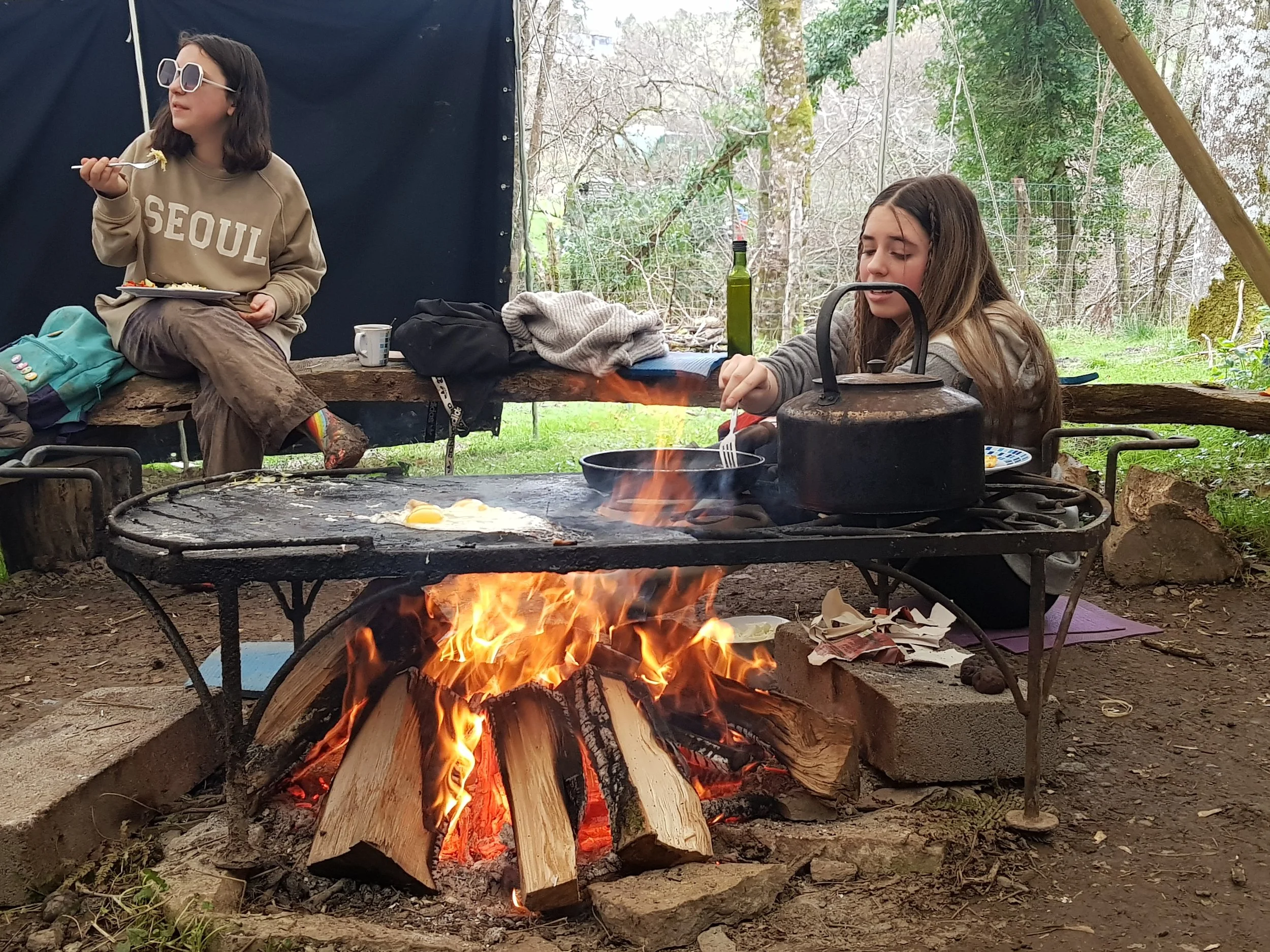 Two young girls sitting outdoors near a campfire. One girl is wearing sunglasses and a sweatshirt, and is eating with a fork. The other girl is focused on poking the fire with a stick. There are trees and a black fabric backdrop behind them, and camping gear on a wooden log.