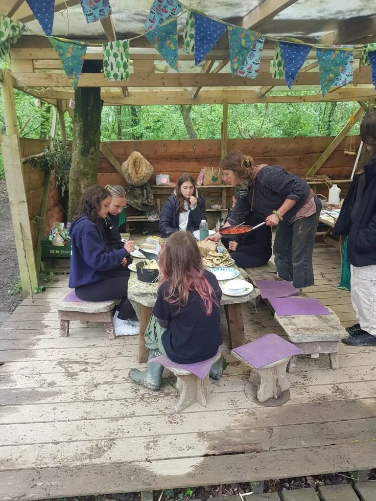 Girls and an adult woman sitting around a low wooden table outdoors, eating food, with a woman serving. The setting is rustic with a wooden deck, makeshift wooden furniture, and decorative bunting flags overhead.