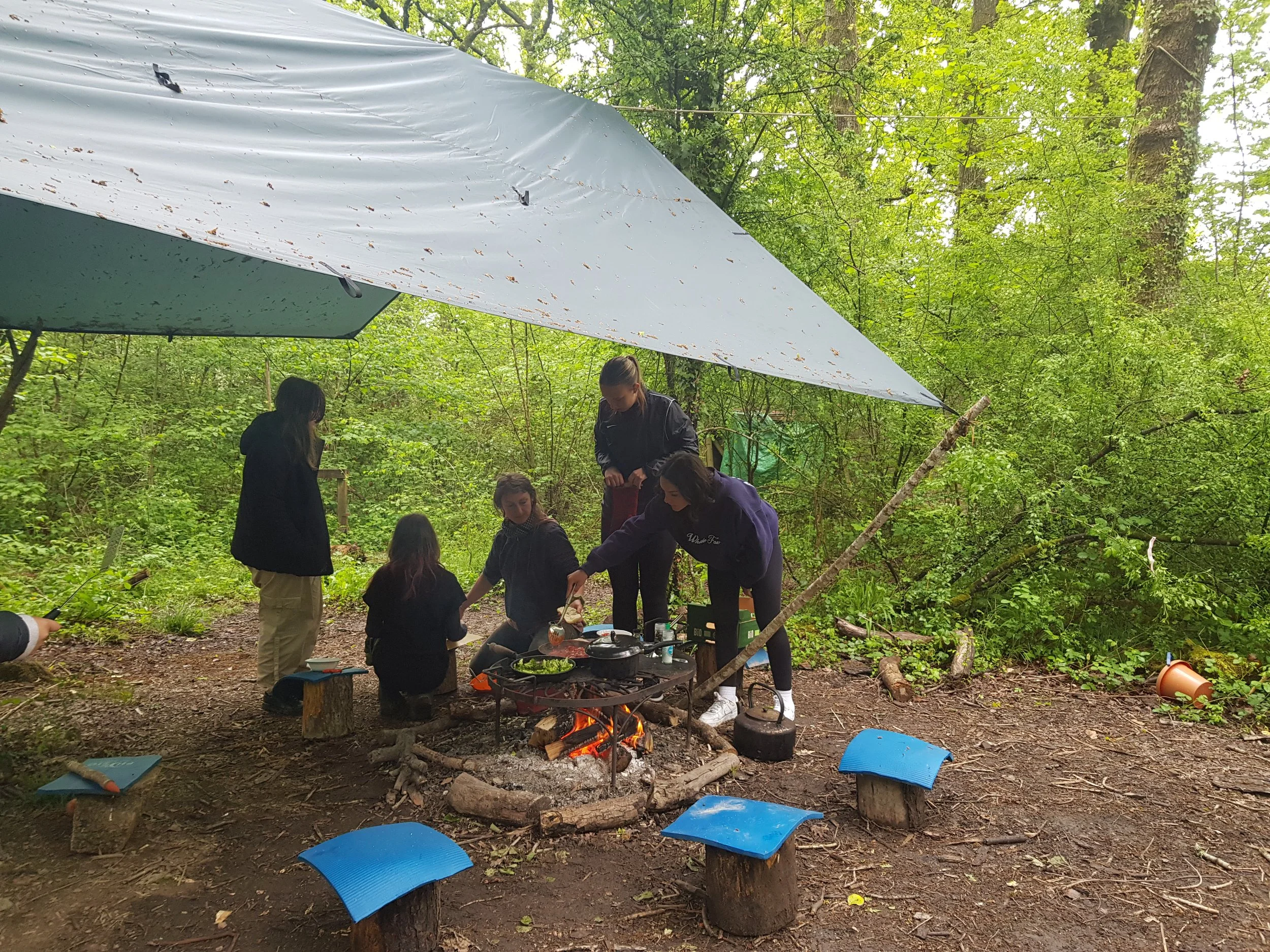 Group of young women gathered around a campfire in a wooded outdoor area, with a tarp overhead, preparing or cooking food.