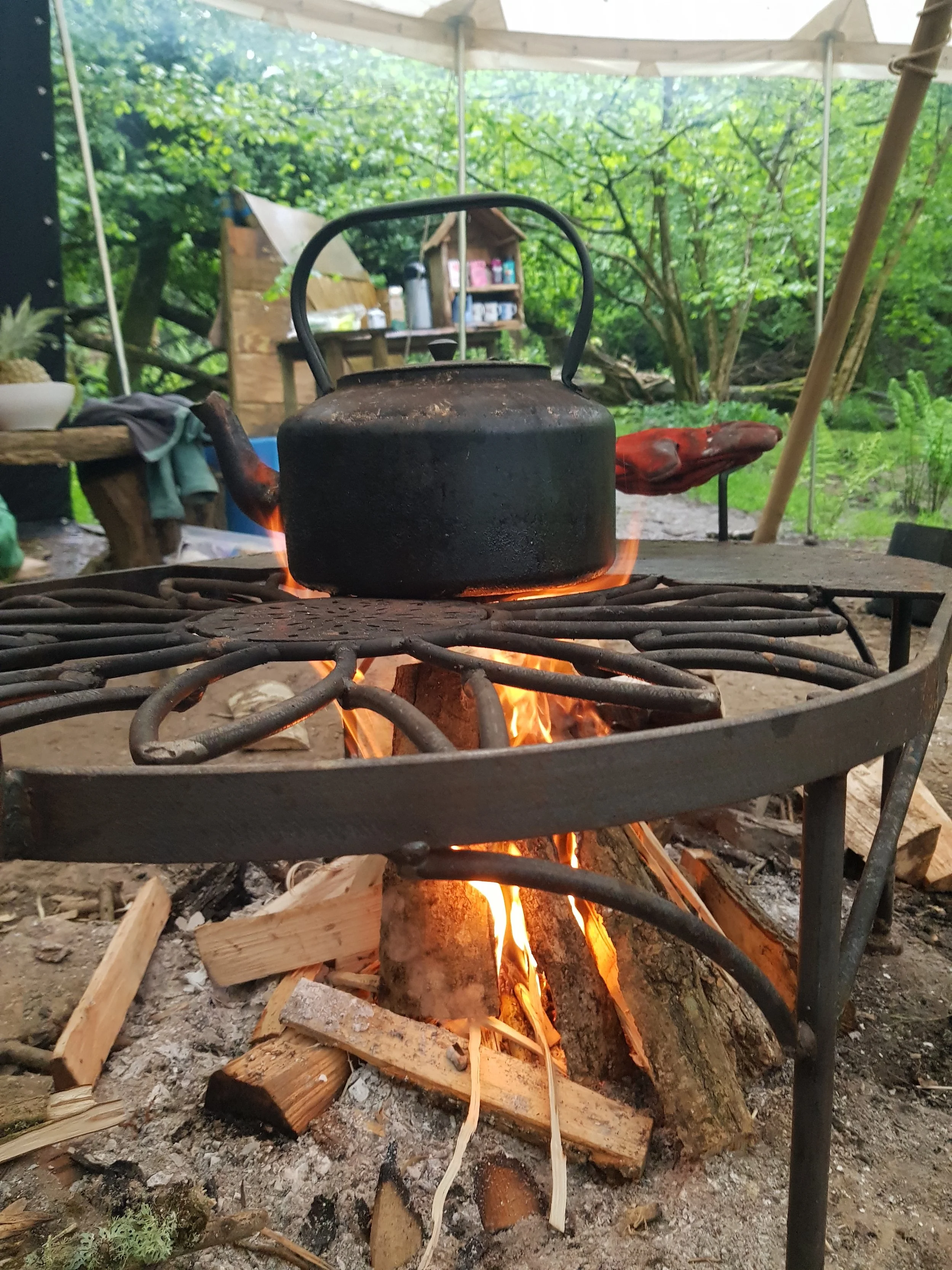 A black kettle on a campfire with wood logs burning below, set outdoors under a canopy with green foliage in the background.