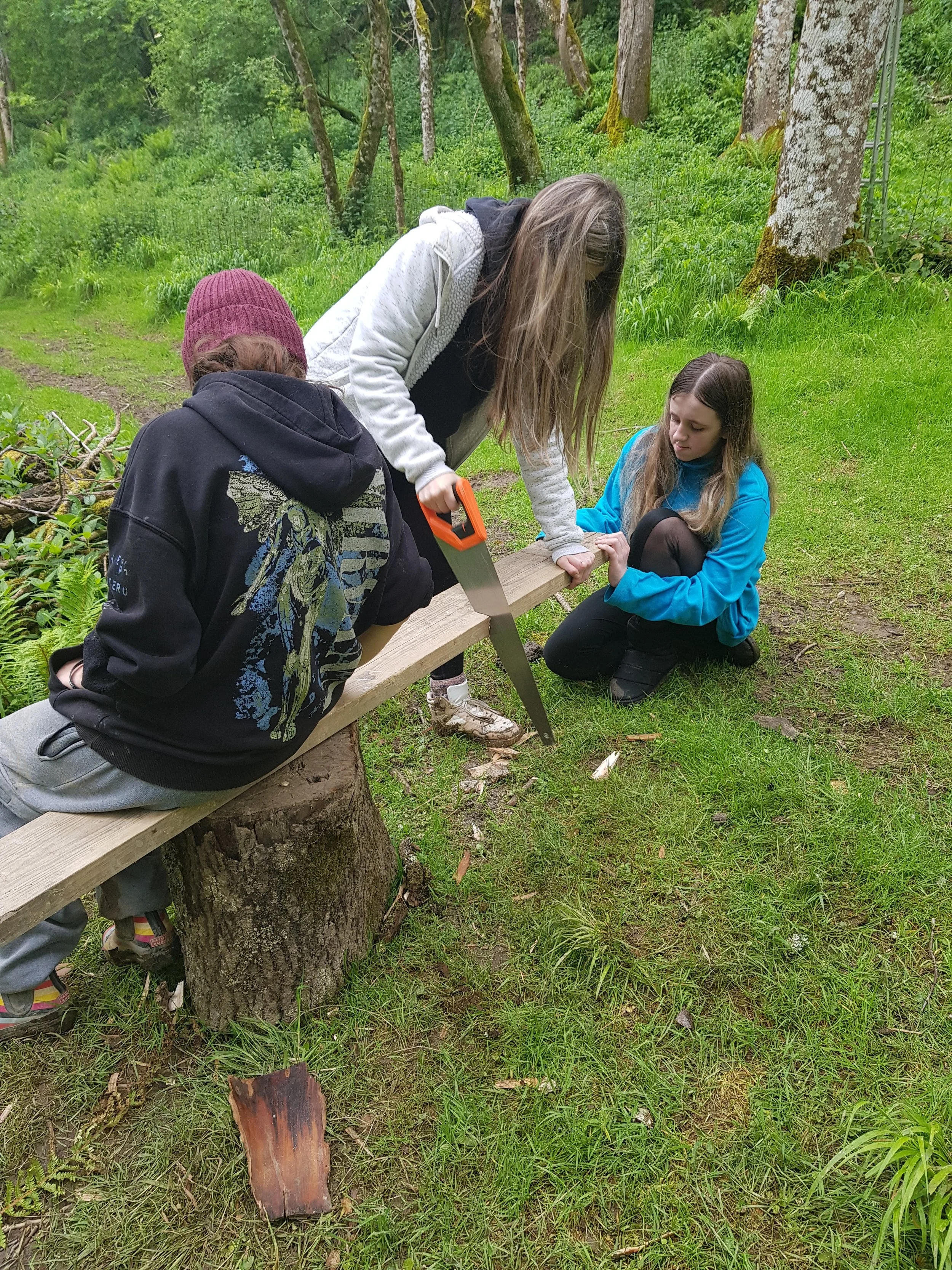 Three young women outdoors in a forested area, working together on a woodworking project. One young woman is sawing a piece of wood while the others observe.