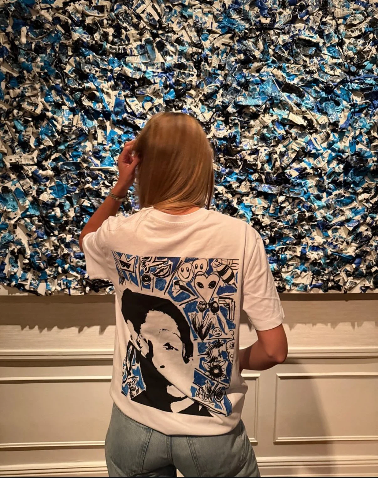 A woman with shoulder-length blonde hair wearing a white T-shirt featuring a black and white portrait of a woman and various blue doodles, looking at an abstract textured wall made of twisted blue, black, and white paper or fabric.