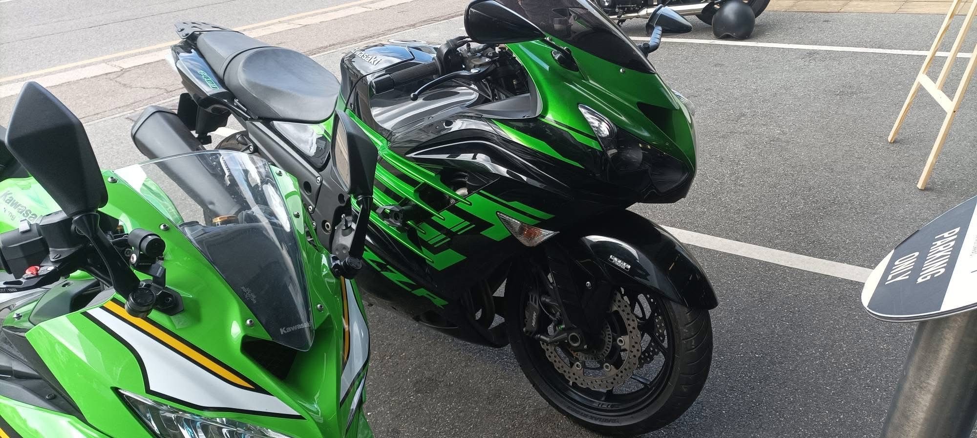 Two green and black Kawasaki motorcycles parked on asphalt in a parking lot, with a 'NO PARKING' sign visible.