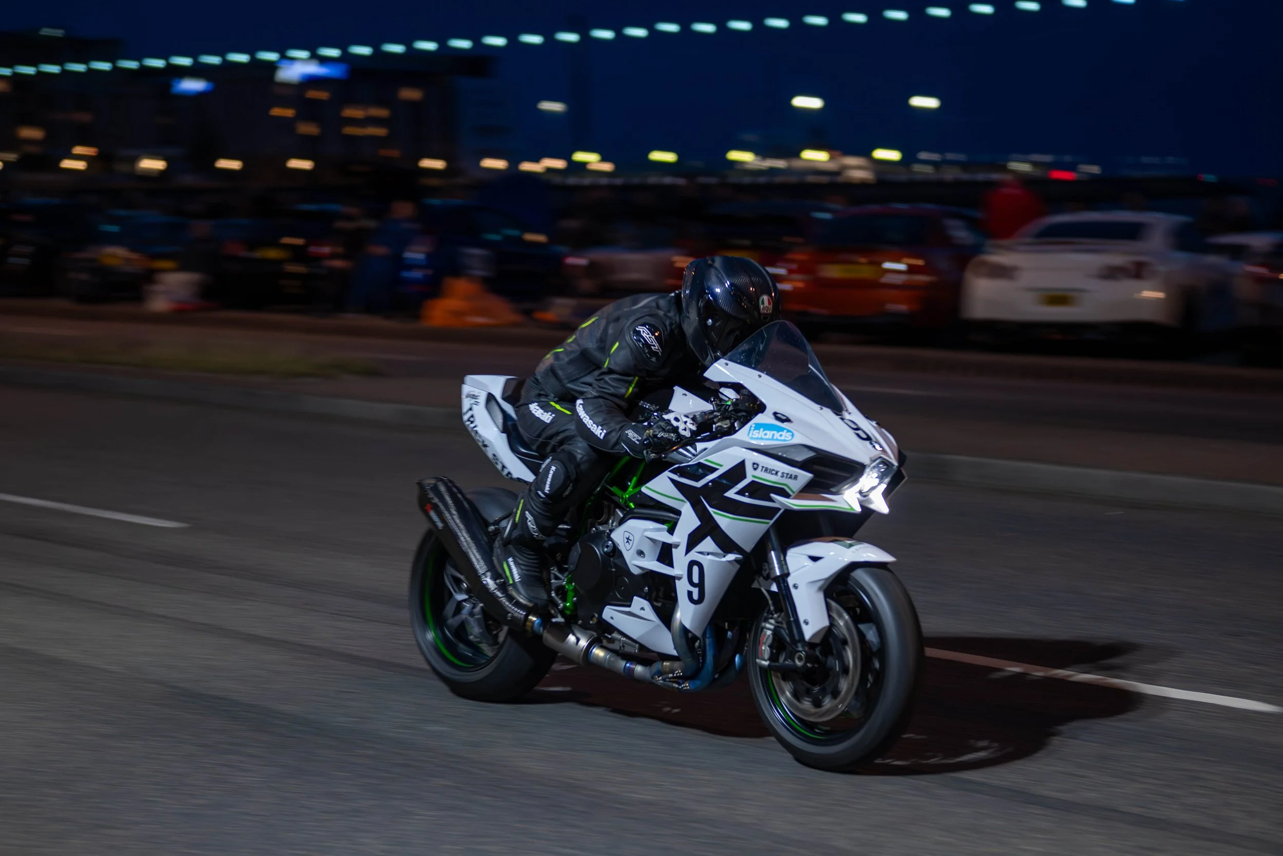A motorcyclist dressed in black riding a white Kawasaki sport bike at night on a city street. The background shows blurred parked cars and city lights.