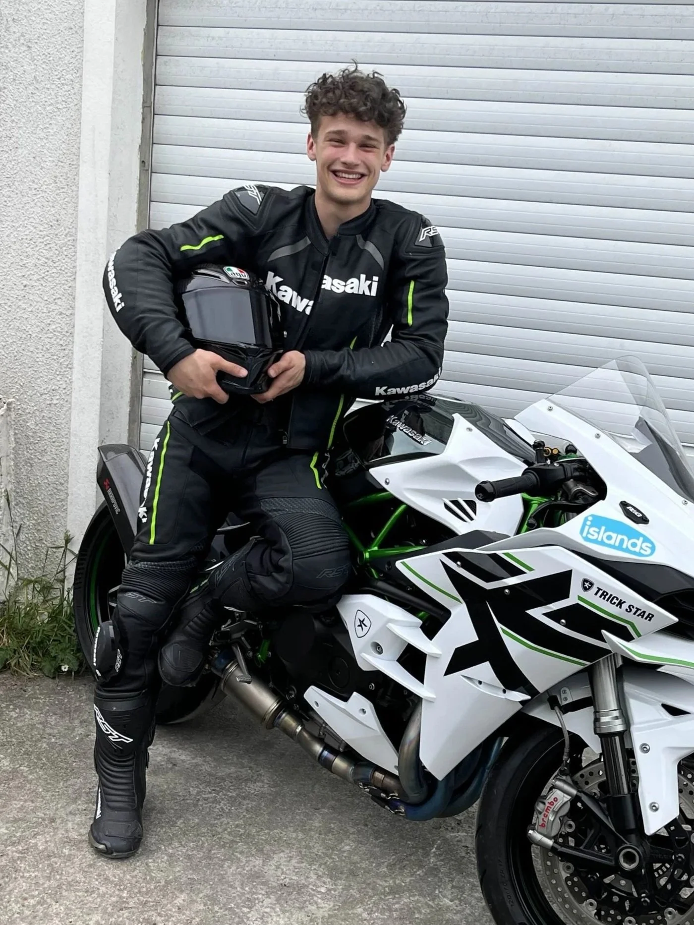 A young man wearing a black motorcycle racing suit, holding a helmet, sitting on a white and green Kawasaki motorcycle, smiling, with a white garage door in the background.
