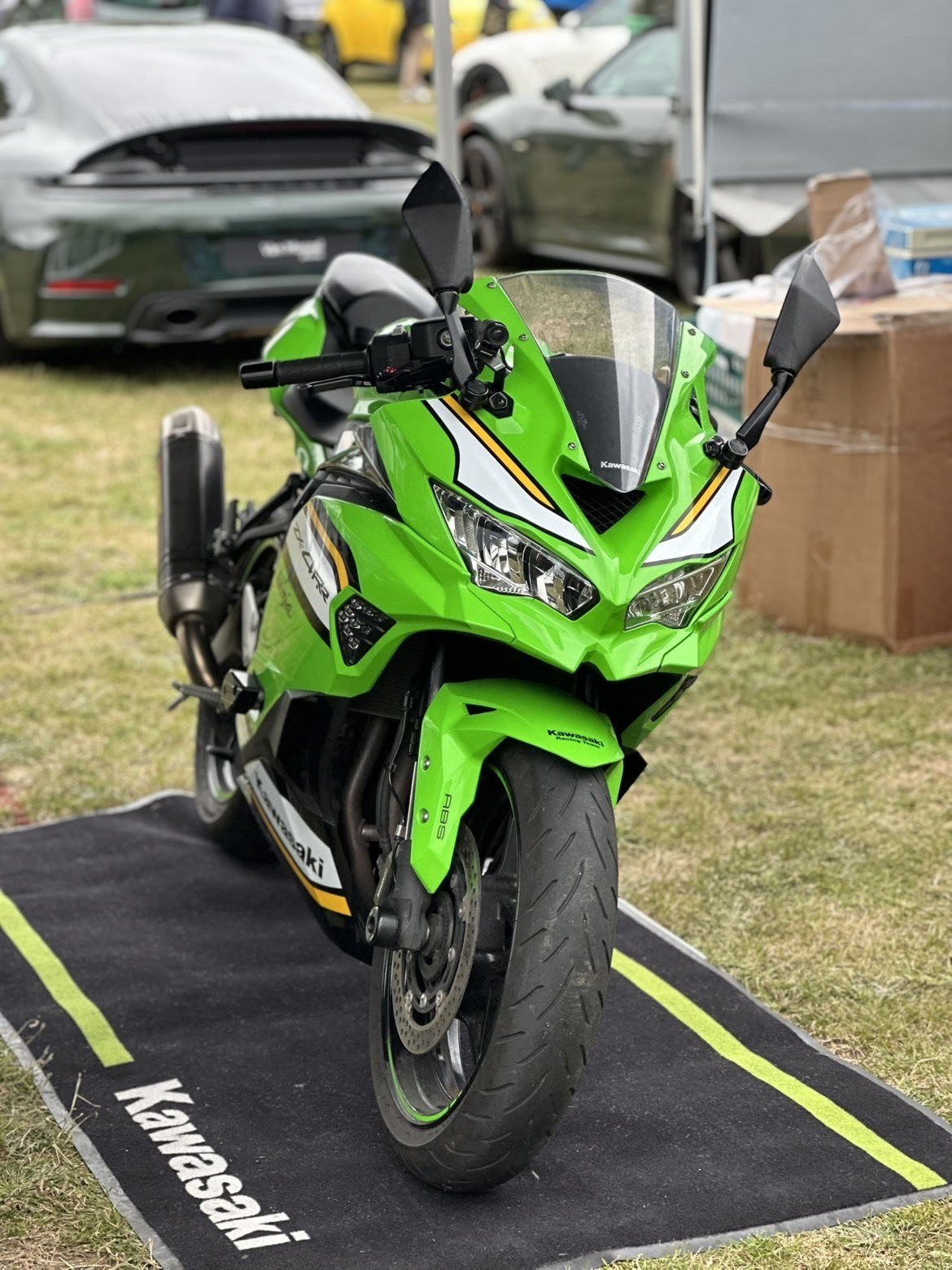 A bright green Kawasaki Ninja sport motorcycle displayed on a black Kawasaki-branded mat outdoors, with several cars visible in the background.