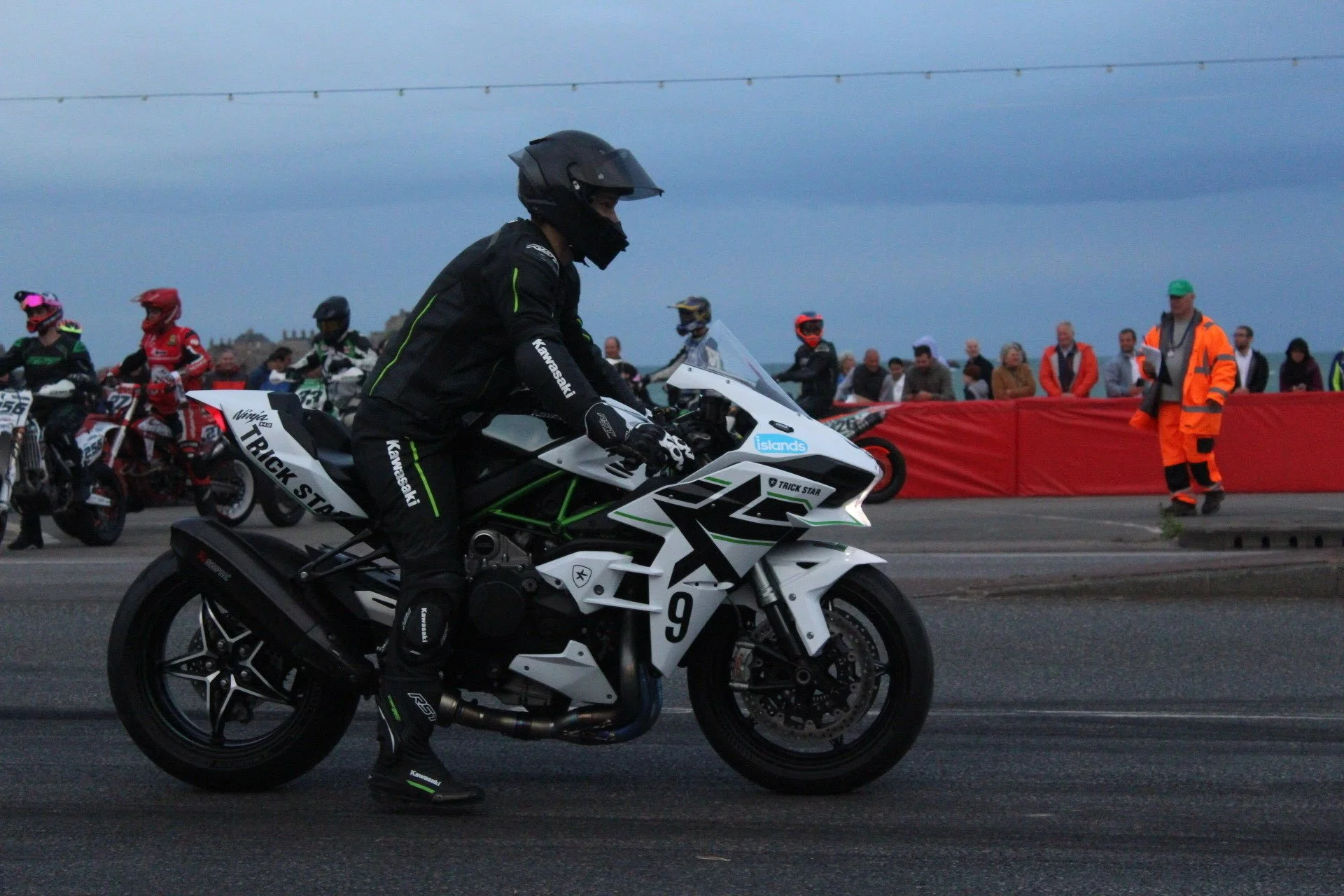 A motorcyclist dressed in black riding a white Kawasaki motorcycle with the number 9, in a race or event setting. There are other motorcyclists and an audience in the background, with some officials in orange and green safety vests.
