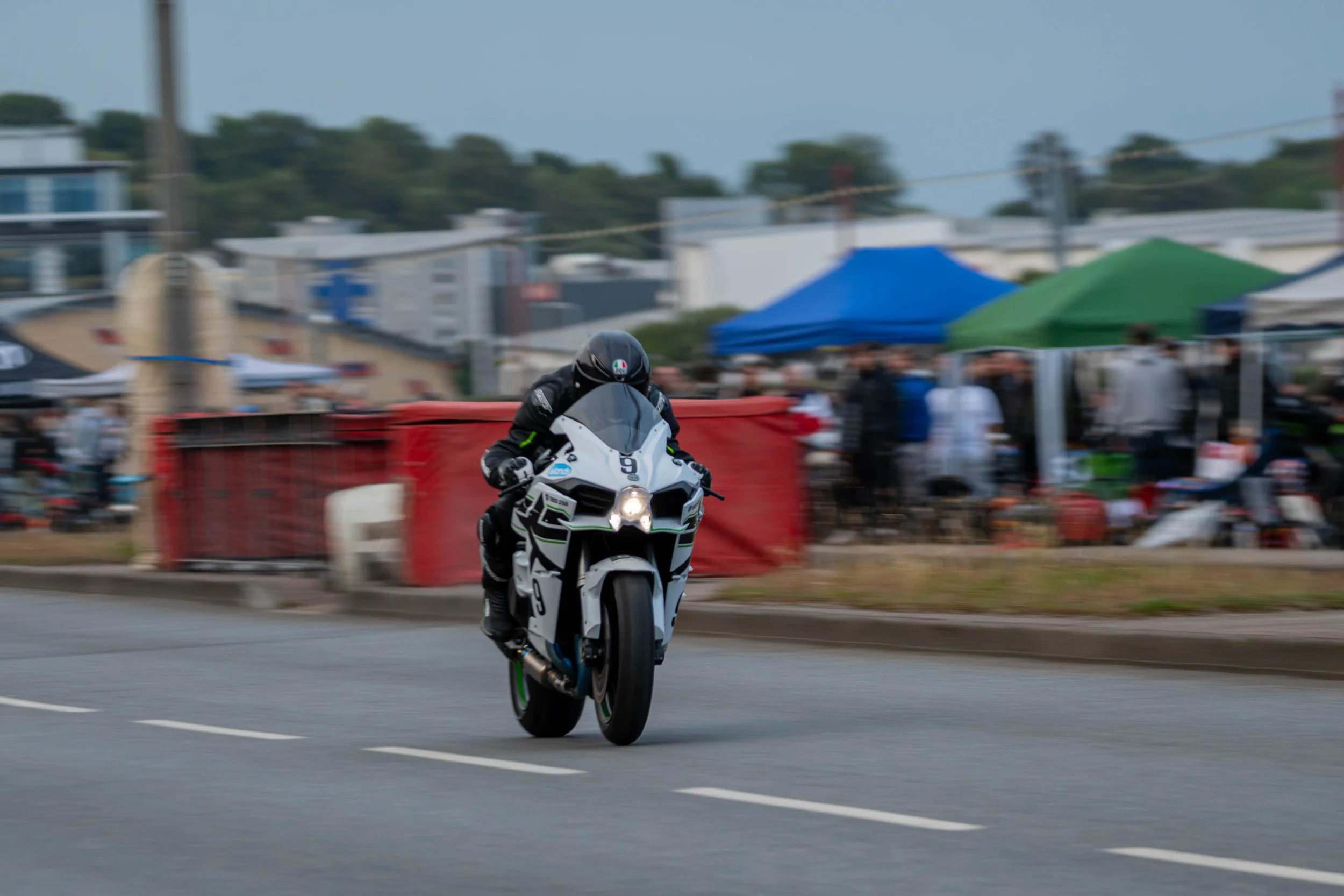 A motorcyclist performing wheelie stunt on a sportbike motorcycle on a street with blurred spectators and tents in the background.