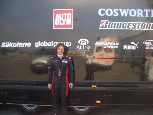 Person in racing suit standing in front of team trailer with various sponsor logos.