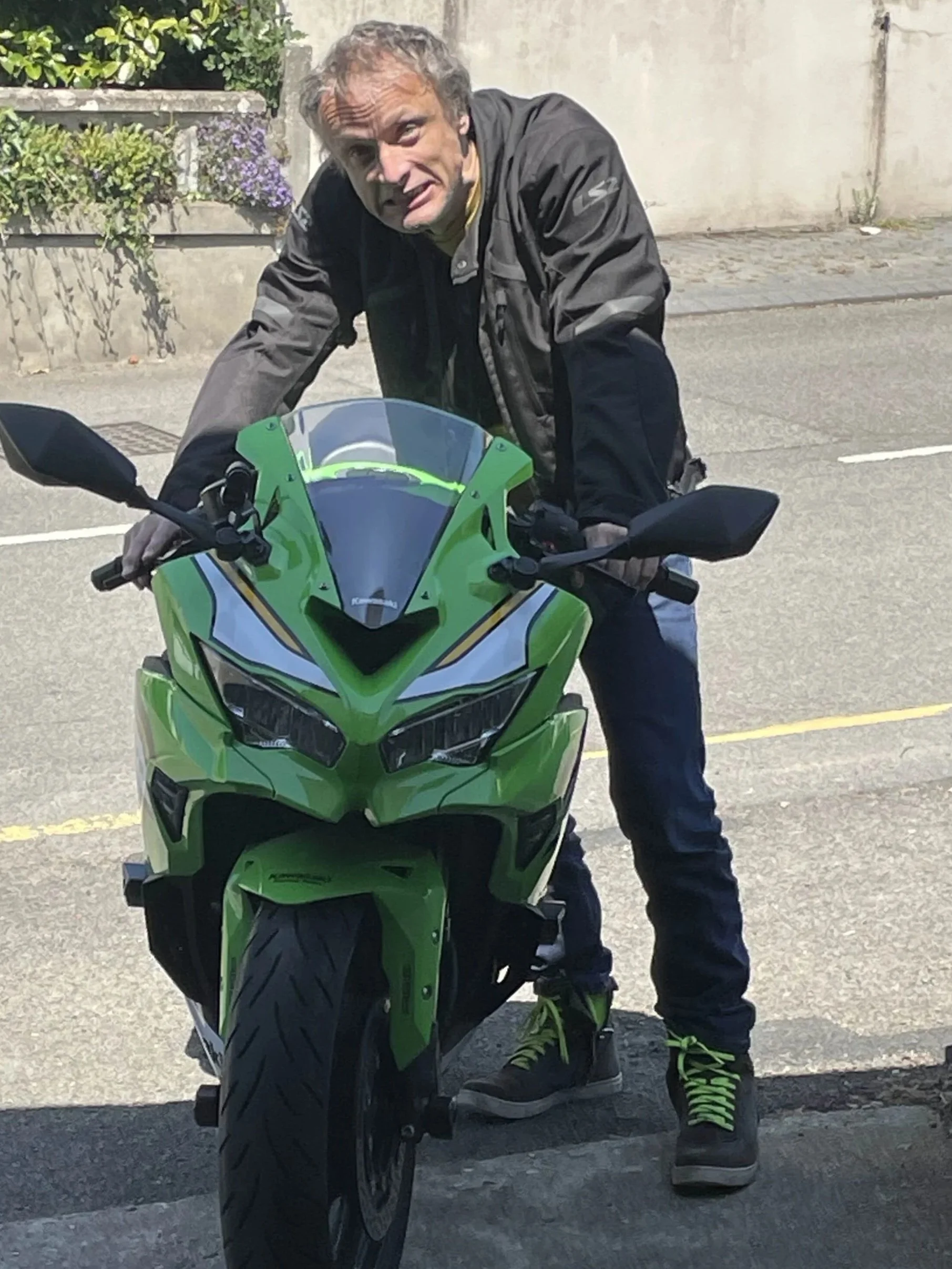 A man with gray hair and a black jacket bent over a green sport motorcycle on a street, with a concrete wall and some plants in the background.