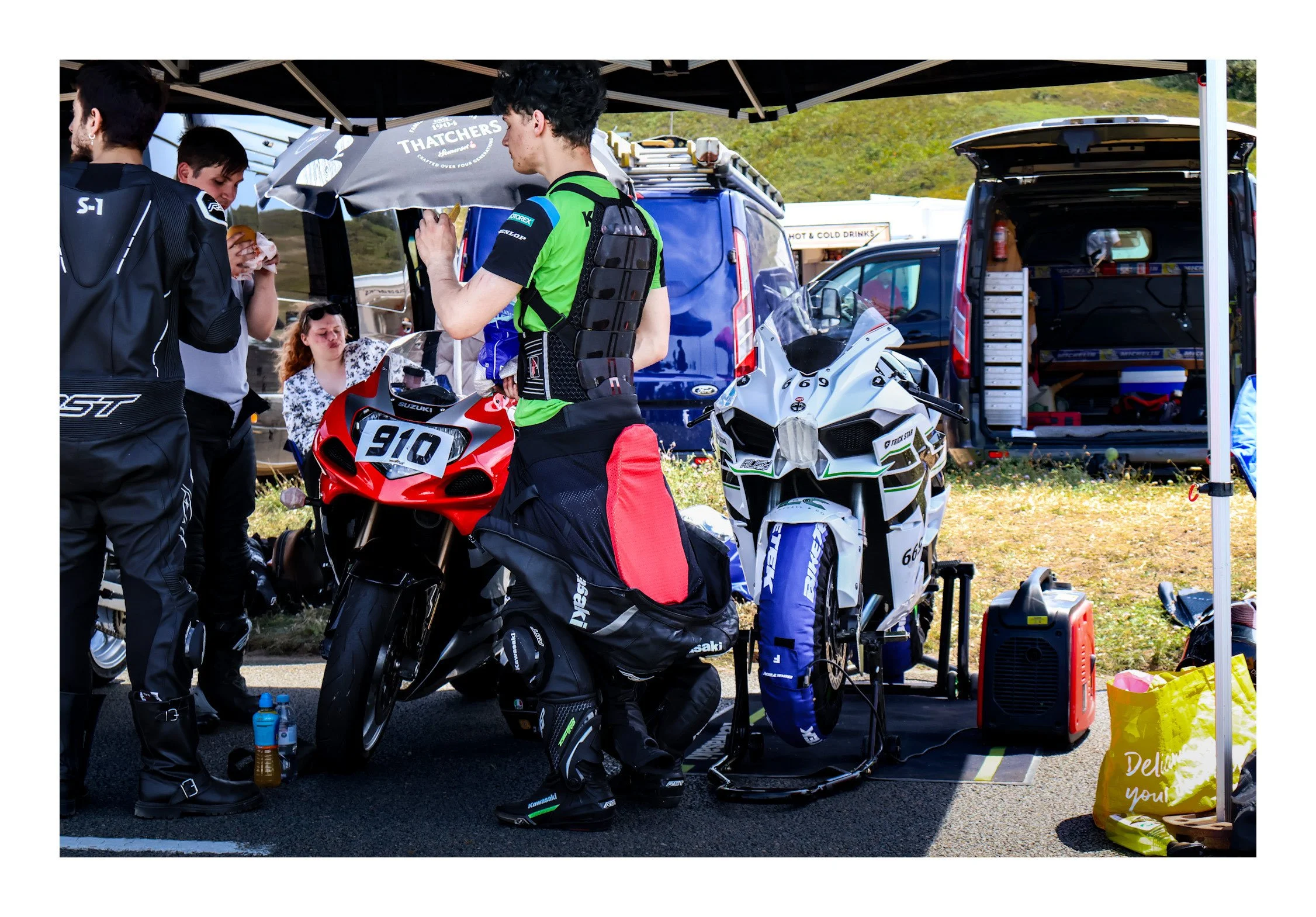 A group of motorcyclists in racing gear gathered at a pit stop or preparation area, surrounded by motorcycles, equipment, and supplies, with a table under a canopy outdoors.