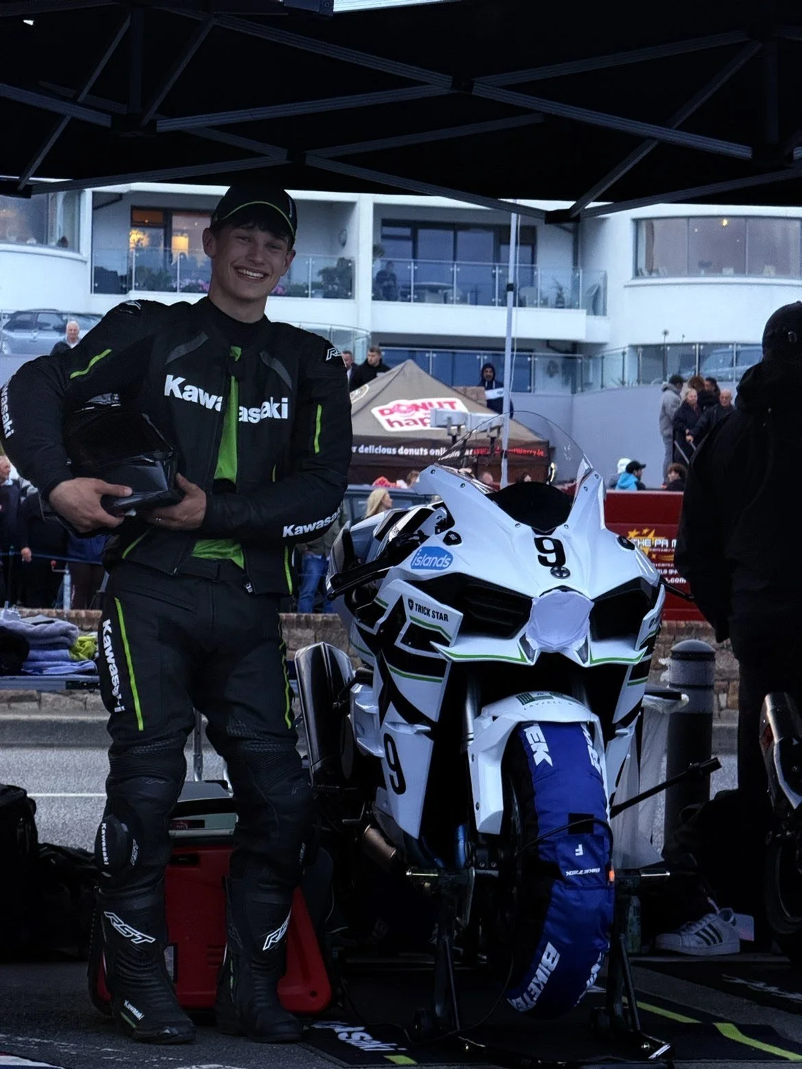 A young man in motorcycle racing gear standing next to a white sport motorcycle, holding a helmet, at a racetrack or pit area during daylight. There are people and buildings in the background.