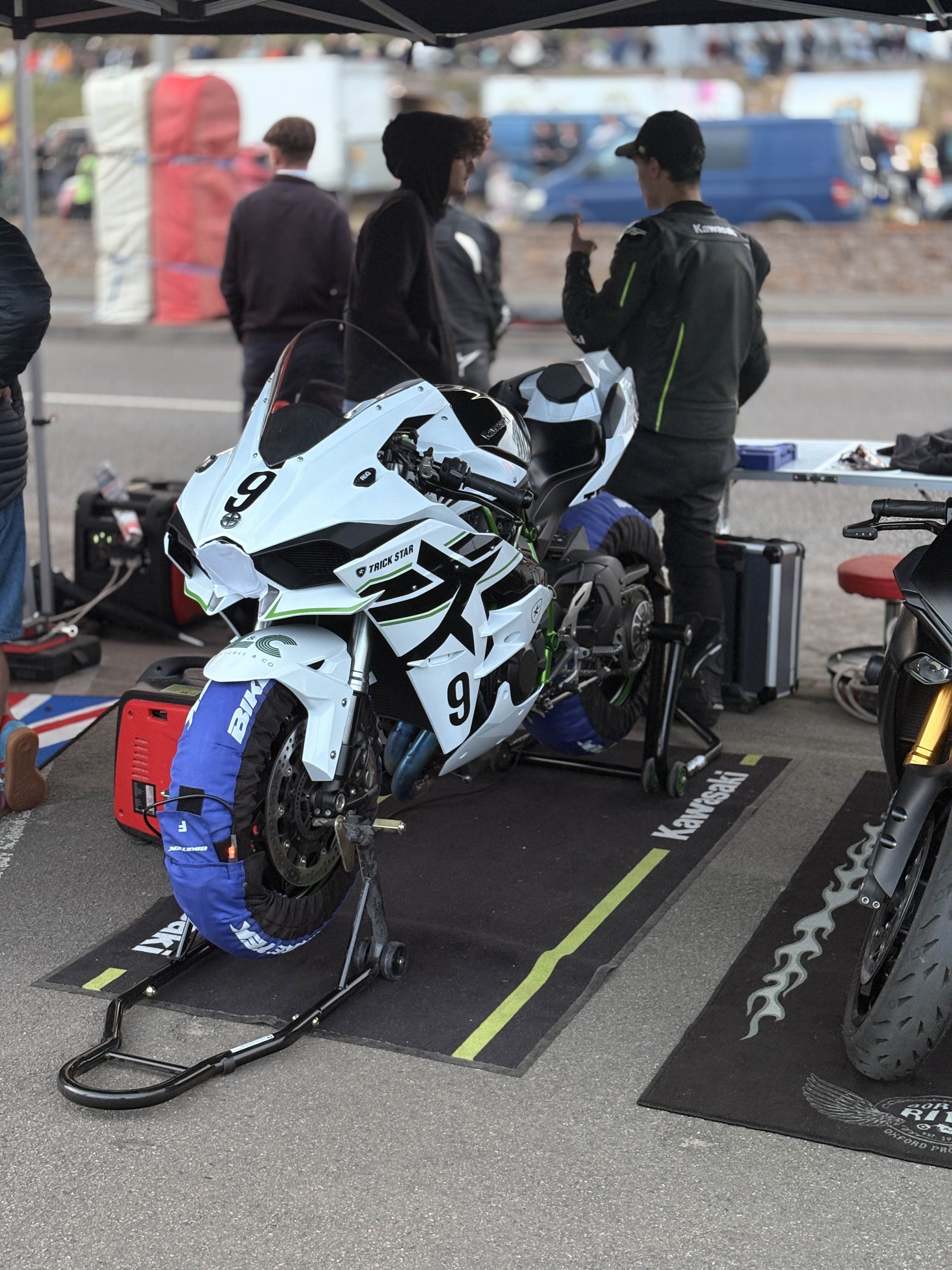 A motorcycle is on a stand in a race team garage, with racing gear and tools around it. Three people are in conversation behind the motorcycle, in a paddock area with parked trucks and trailers in the background.