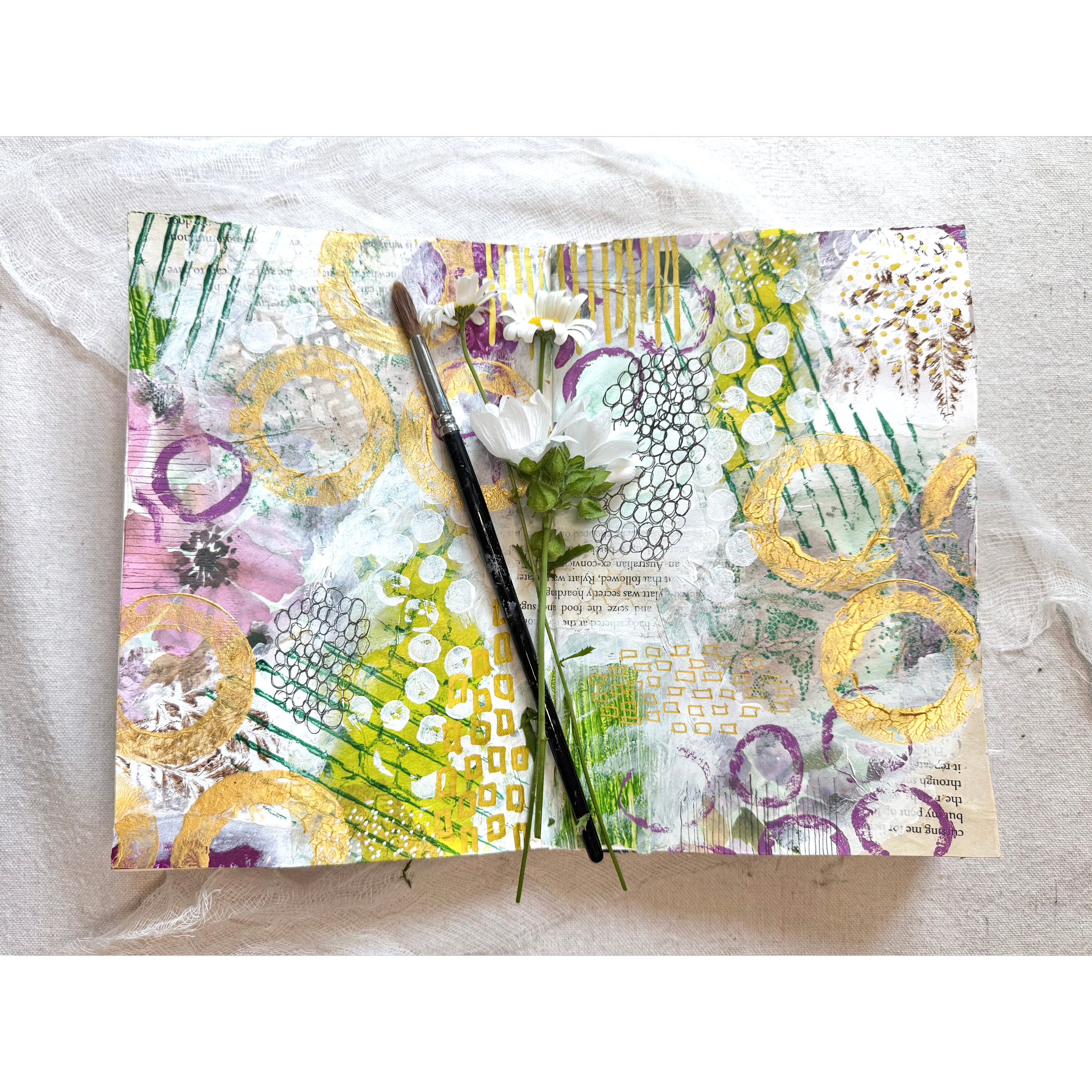 Mixed media art journal page with abstract colorful patterns, gold accents, white flowers, and a paintbrush on textured fabric background