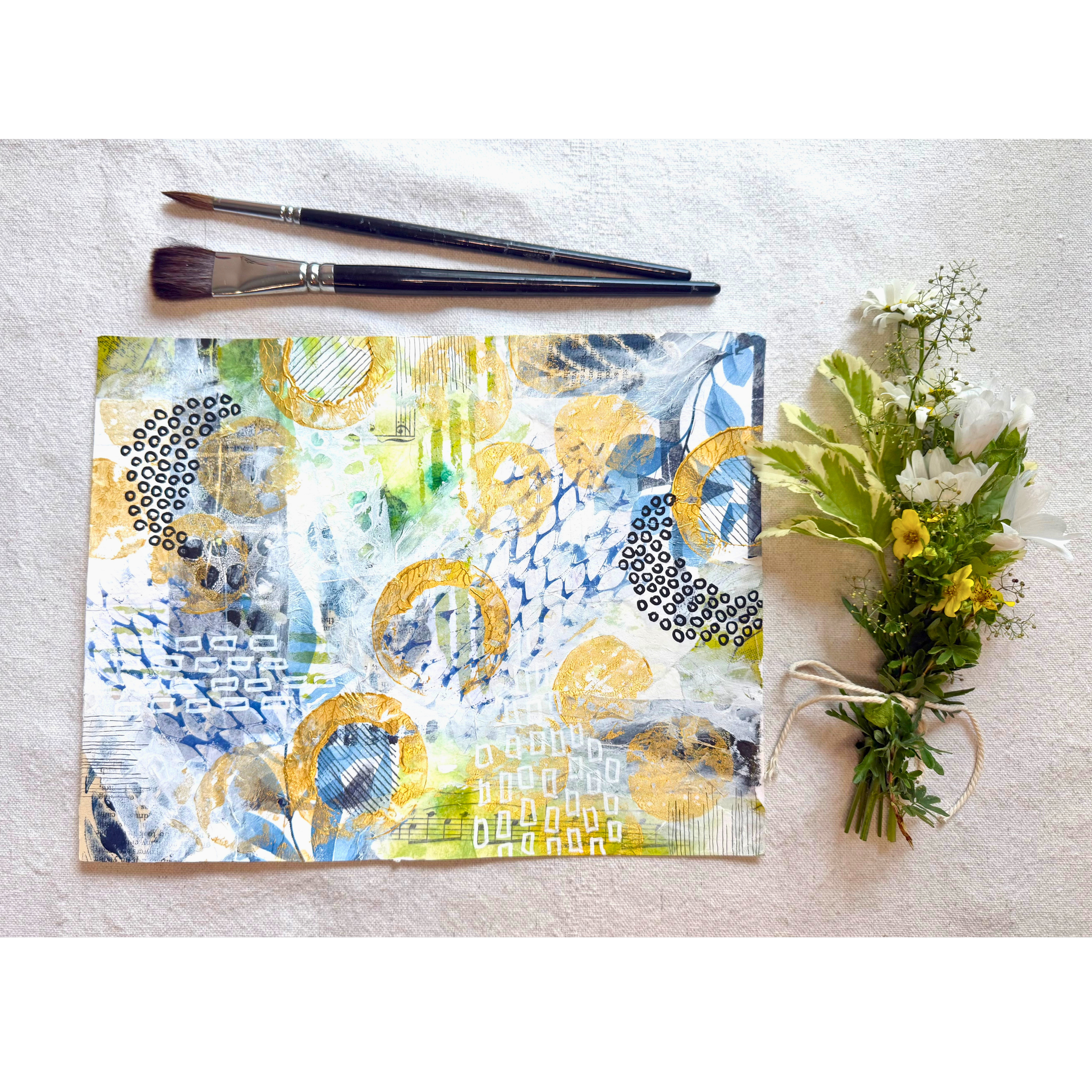A paintbrush and a collage art piece with a small bouquet of white and yellow flowers on the side.