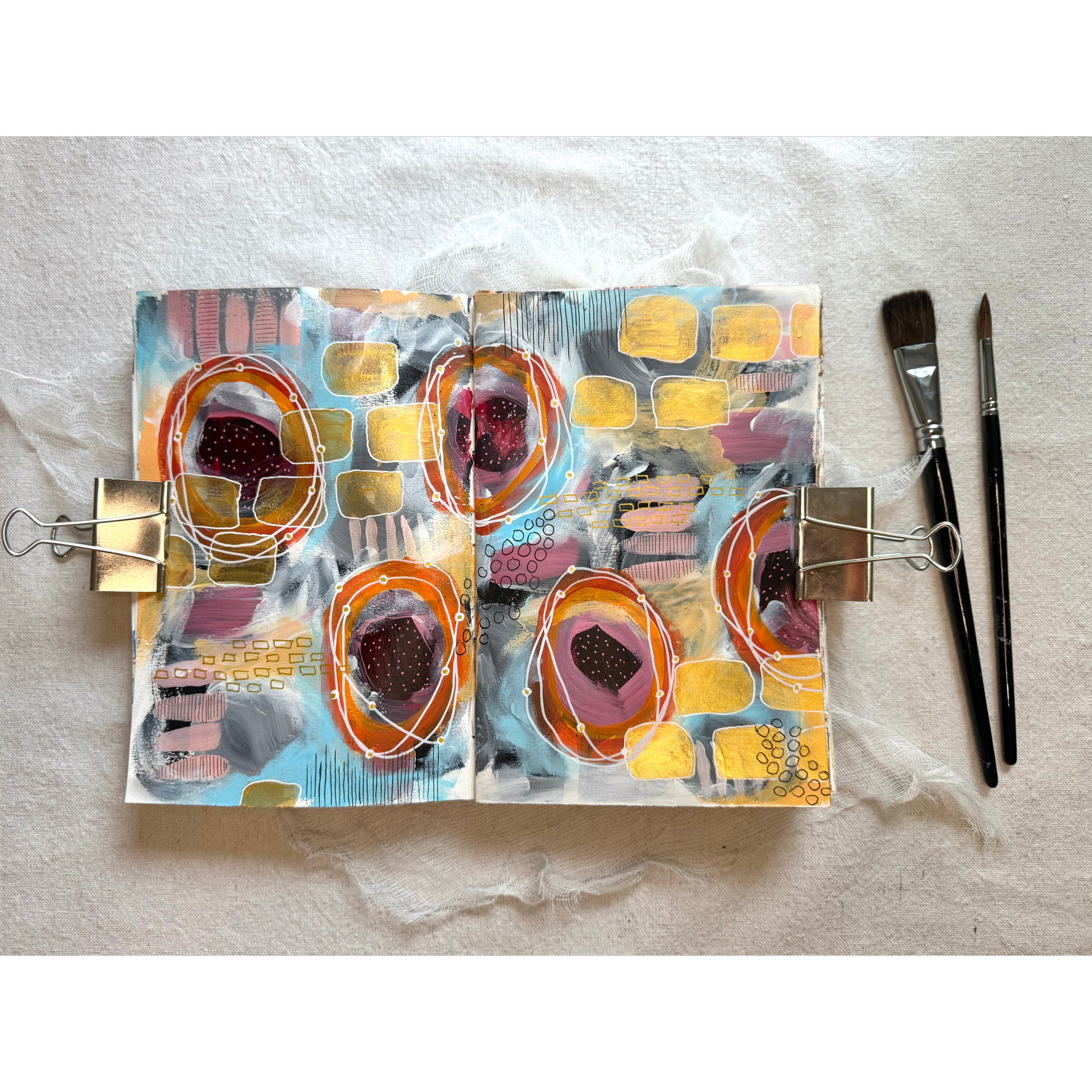 Open sketchbook with colorful abstract art featuring circles, squares, and lines, placed on a white textured surface with two paintbrushes beside it.