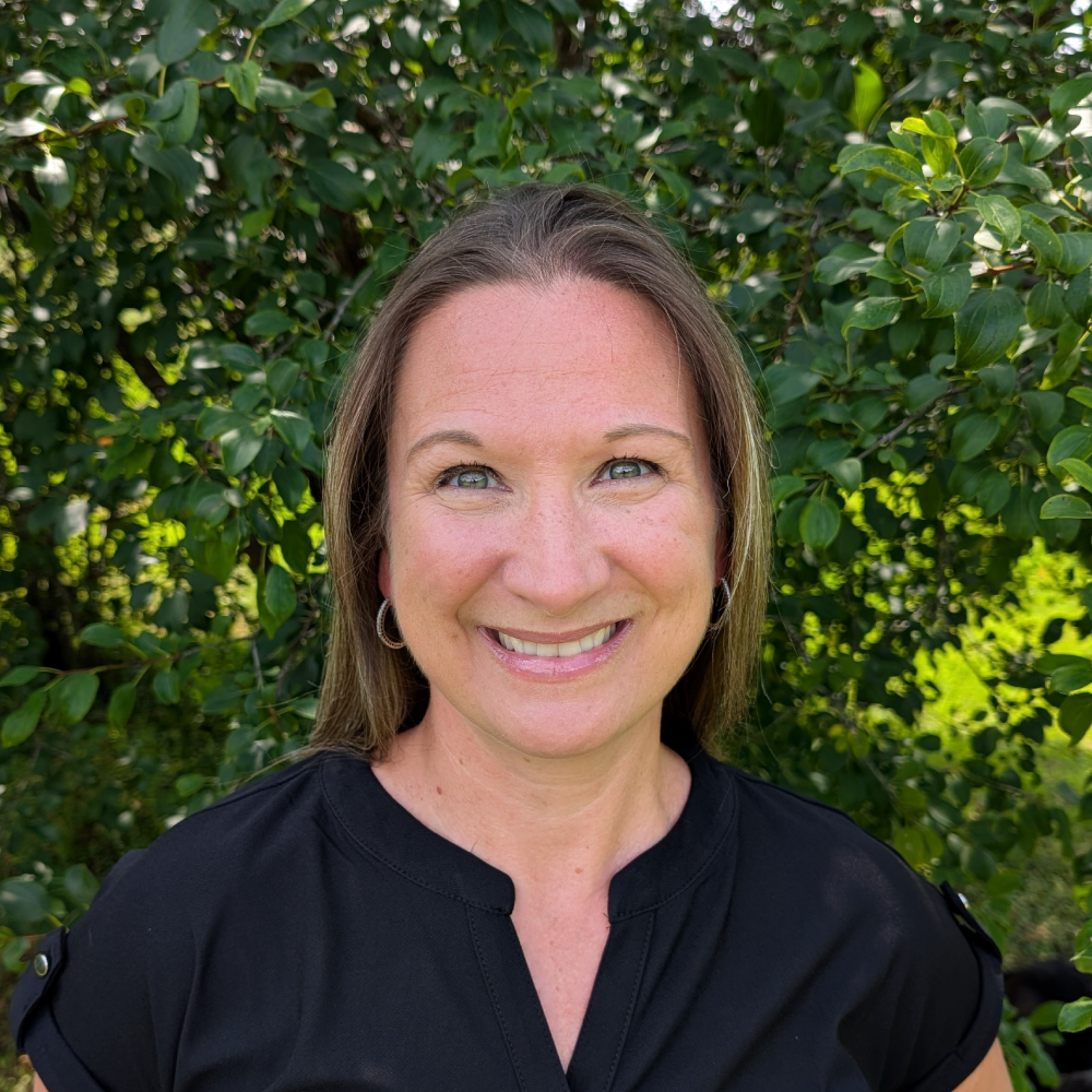 A woman smiling outdoors in front of green leafy bushes, wearing a black top.