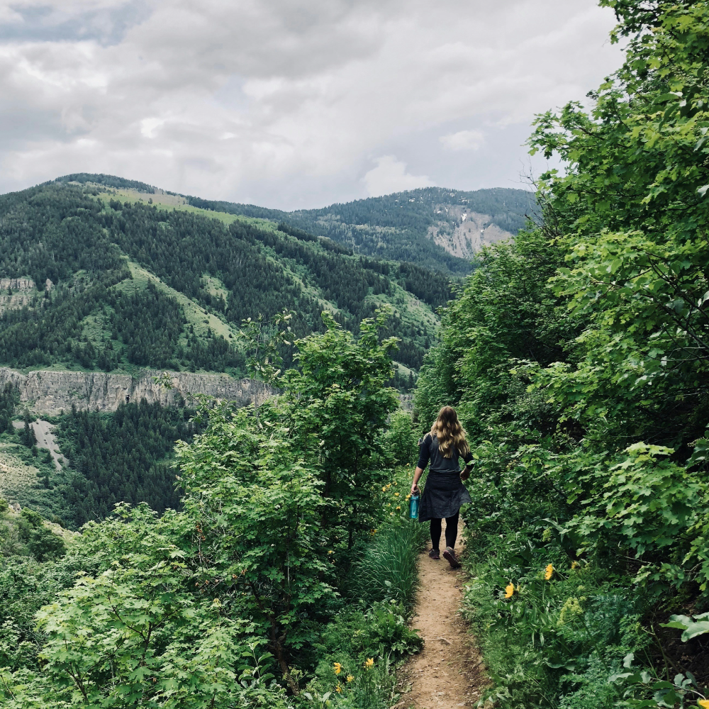A person with long hair, dressed in dark clothing, walking along a narrow dirt trail surrounded by lush green trees and shrubs, with a backdrop of rolling green mountains under a cloudy sky.