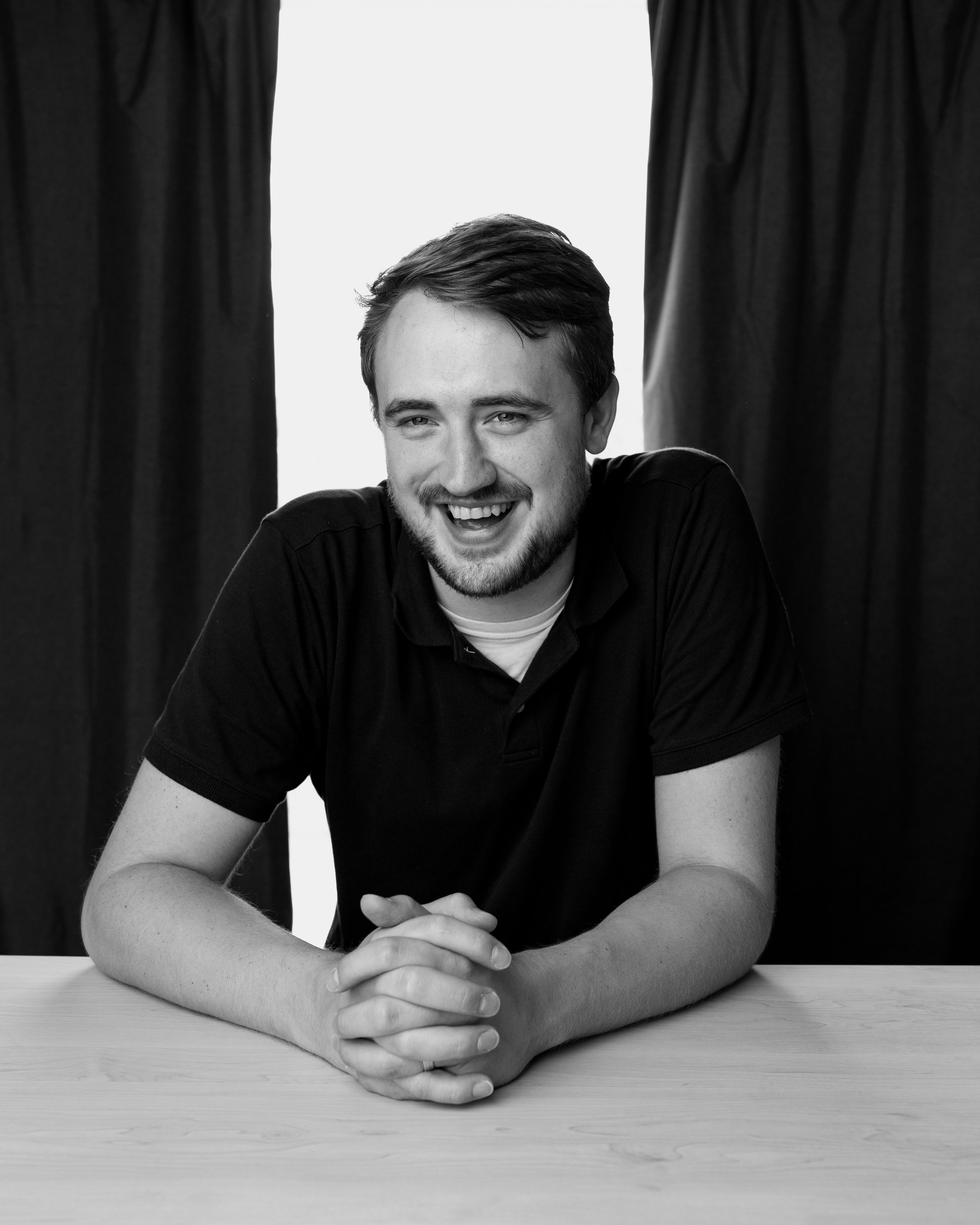 A young man with brown hair and a beard, smiling and sitting at a table with his arms resting on it, hands clasped together. The image is in black and white, with dark curtains behind him and a plain white background.
