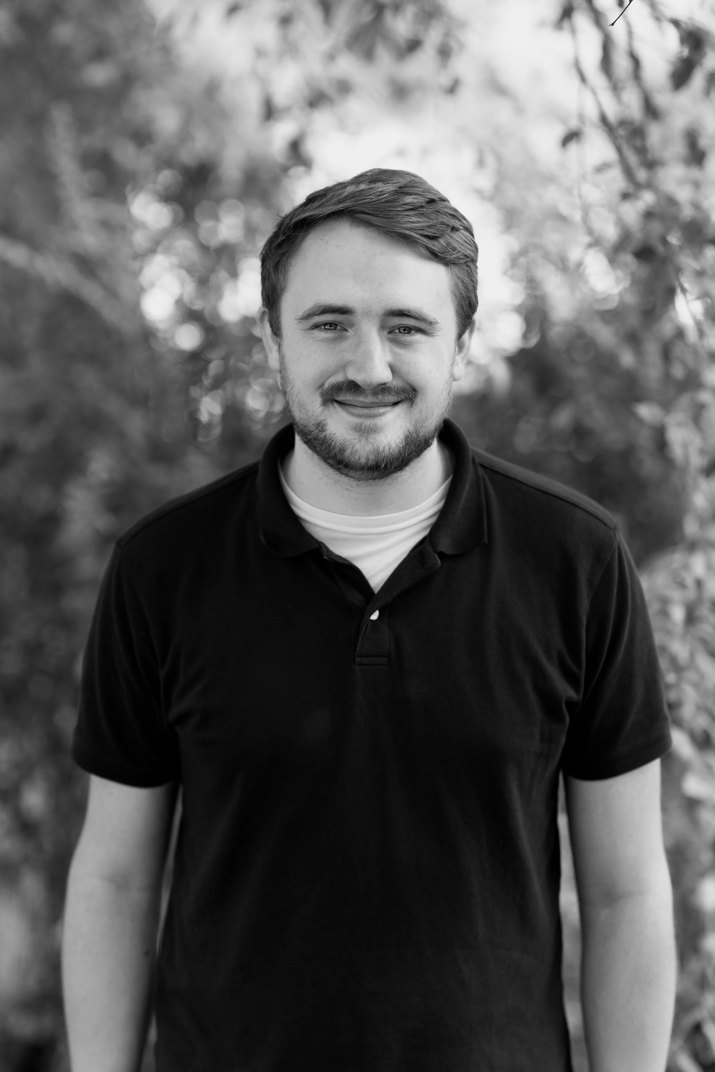 Black and white portrait of a young man with a beard and short hair, smiling, wearing a black polo shirt, standing outdoors with blurred trees in the background.