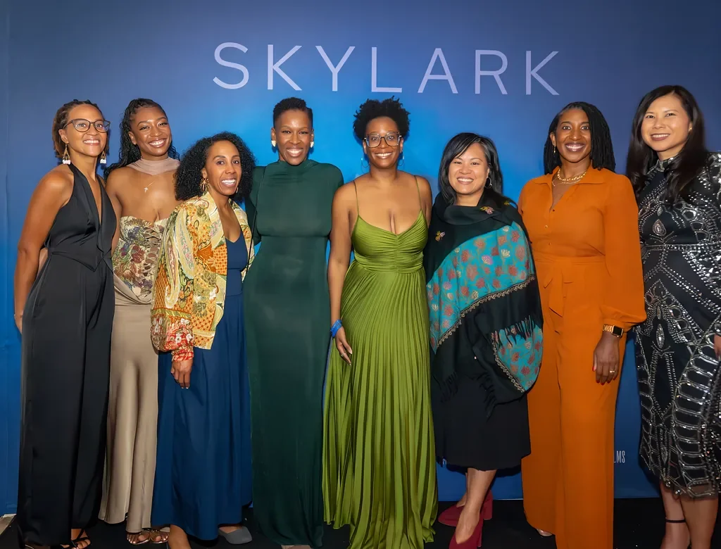 A group of eight women smiling and posing together in front of a blue background with the word 'SKYLARK' displayed at the top.