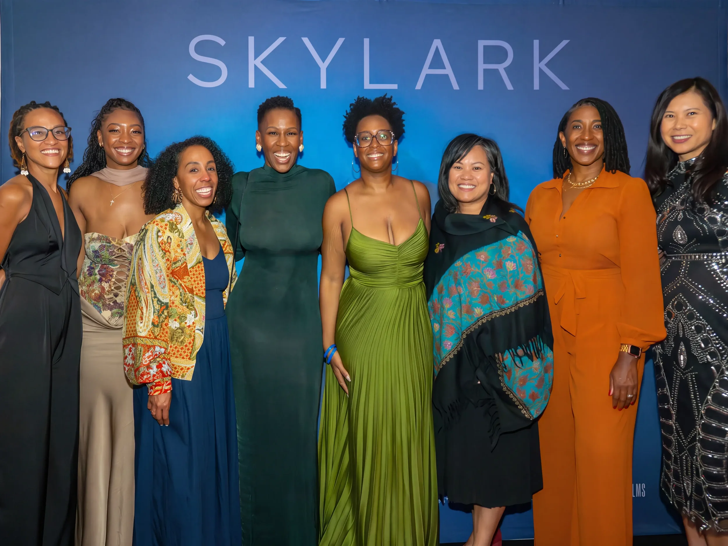 A group of eight women smiling and posing together in front of a blue background with the word 'SKYLARK' displayed at the top.
