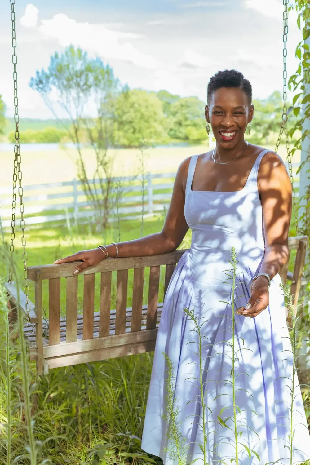 A woman in a white dress stands outdoors next to a porch swing, smiling at the camera, with a lush green background and shadows from trees.