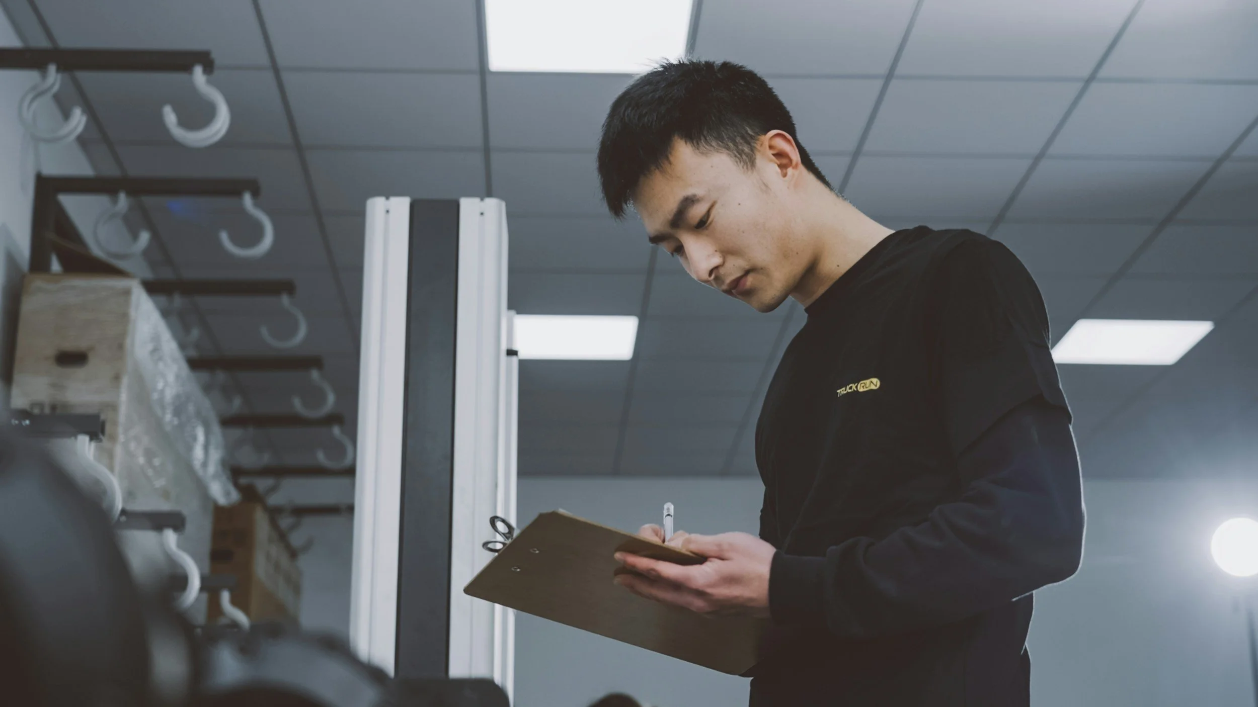 A young man with black hair and a black shirt writing on a clipboard in a gym or workout space with pegs and a wooden box visible in the background.