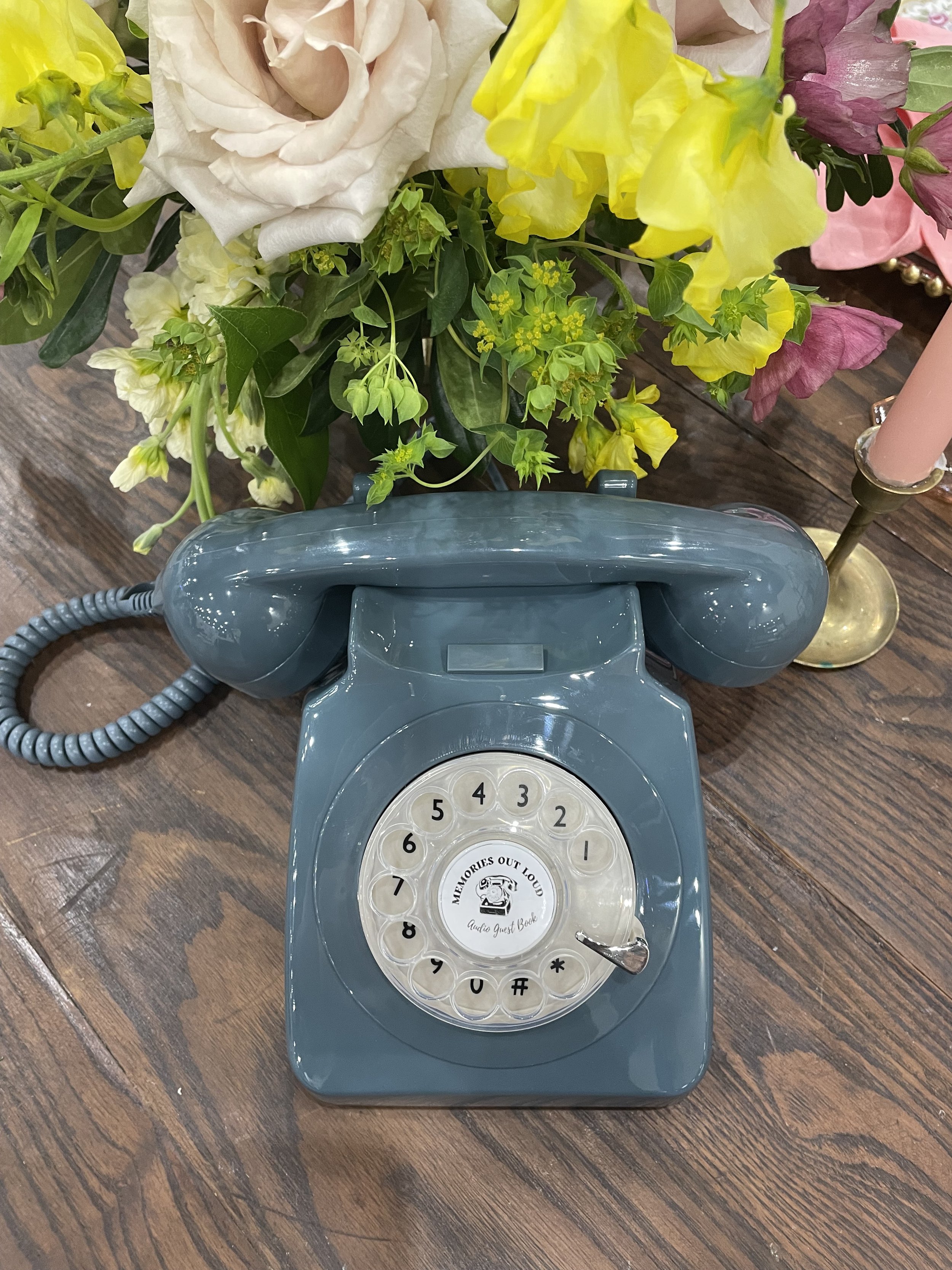 A slate blue rotary audio guestbook phone by Memories Out Loud displayed on a dark wood table alongside colorful yellow and blush floral arrangements.