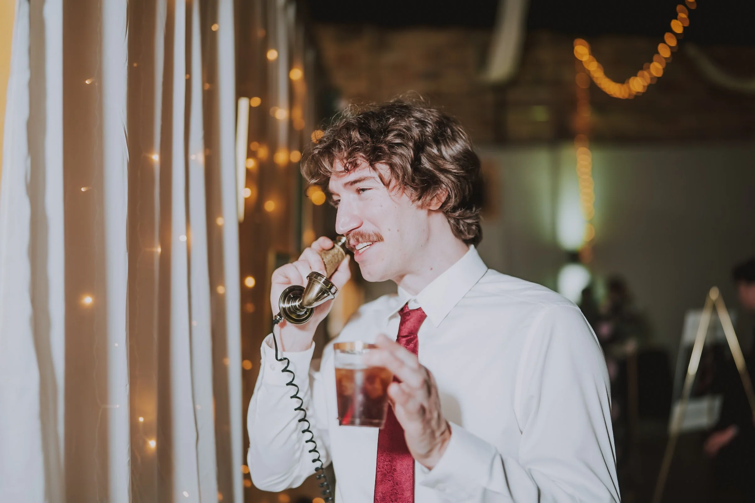 A wedding guest in a shirt and tie holding a drink while leaving a voice message on an antique brass Memories Out Loud audio guestbook phone.